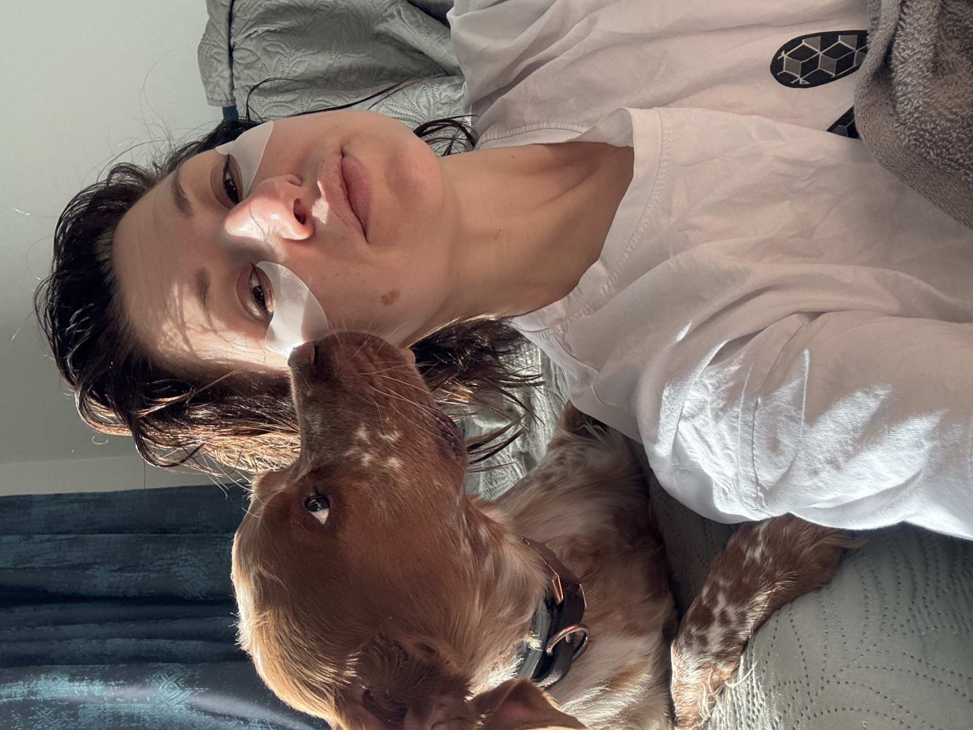 A woman wearing white eye mask patches lies on a light blue patterned bedspread with a tan and white speckled dog, possibly a spaniel, its nose near her face. She wears a white t-shirt and dark hair, and a gray blanket is partially visible. The background is partially visible, with a blue curtain. The location is unknown.