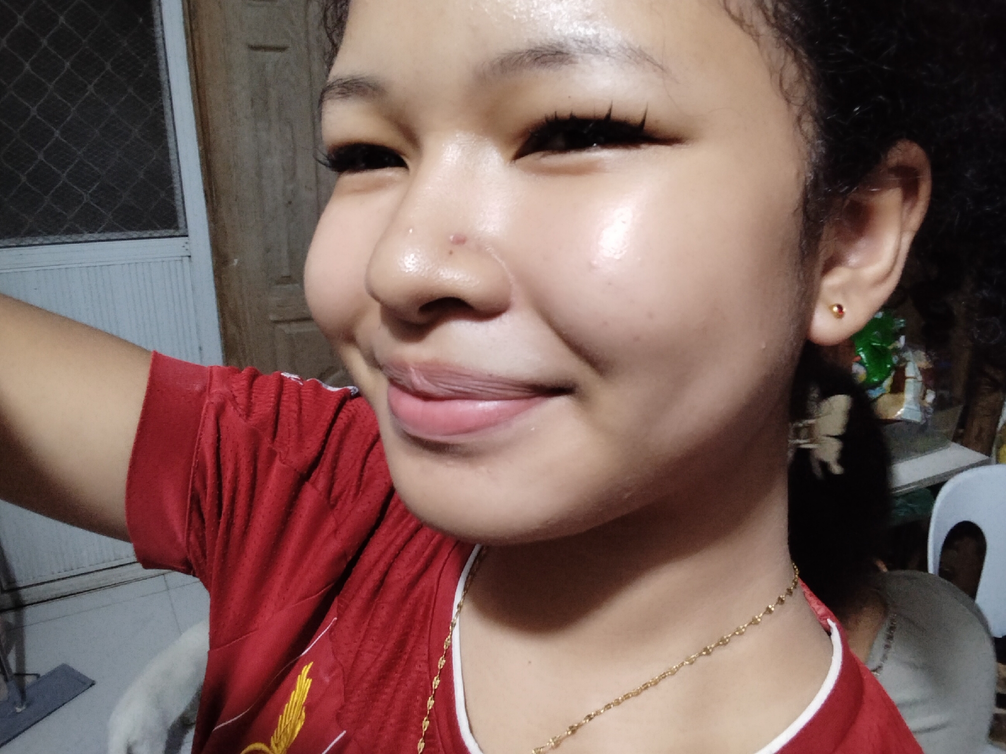 This image is a close-up, selfie-style portrait of a young person in what appears to be a residential setting. The person, who is positioned to the right of the frame and looking towards the camera, has dark, curly hair. They are wearing a red shirt with a white trim around the neckline and a gold necklace. The person's face is illuminated, highlighting their features including applied makeup on their eyes and lips. There is a small red mark on their nose. In the background, to the left, a door with a latticed window is visible. To the right, shelves with various items are partially obscured by the person's hair and a white chair. The lighting suggests an indoor environment, possibly at night. The description does not contain any specific information that would allow for localization to Davao City, Philippines.