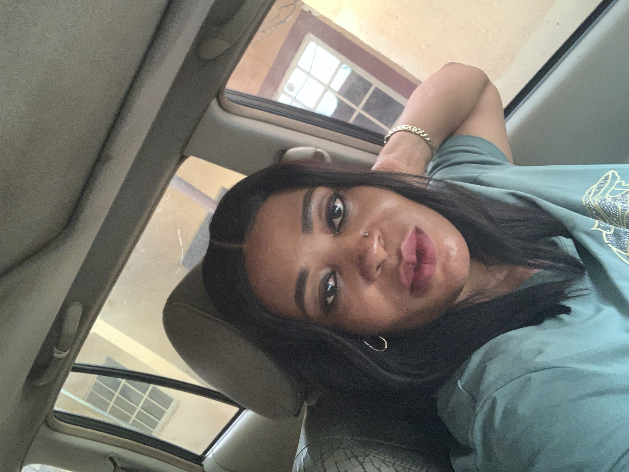 A woman with dark hair and a nose piercing is reclining in a car seat. The car has an open sunroof, and a building with a window is visible through the sunroof. She is wearing a green shirt and a bracelet. This image was taken in Sabon Tasha, Nigeria.