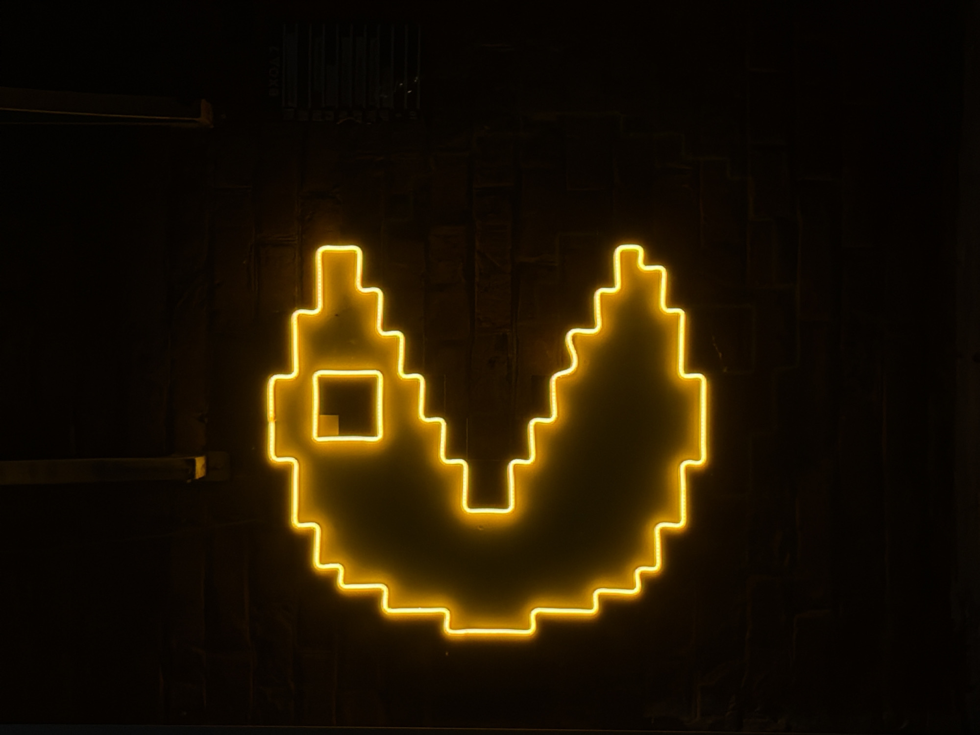 The image displays a brightly glowing, yellow neon sign, shaped like a pixelated rendition of the iconic Pac-Man video game character, set against a dark, textured background. The sign exhibits a distinct blocky, stepped outline, reminiscent of 8-bit graphics, and features a small square cutout towards its upper left, resembling an eye or a missing pixel.
The warm, radiant yellow light of the neon tubing vividly illuminates the character, creating a strong contrast with the predominantly dark and subtly textured wall behind it, which appears to be made of dark material like brick or concrete. The overall darkness of the scene suggests either a nighttime setting or an indoor environment with low ambient lighting, allowing the neon glow to be the primary light source and focal point.
In the upper left corner of the frame, faint details of what appear to be industrial elements, possibly shelving, pipes, or vertical bars (perhaps part of a grate or railing), are subtly visible against the dark backdrop. These elements hint at an urban, industrial, or entertainment venue setting, such as an arcade, bar, or themed establishment. There are no people visible in the image, nor is there any discernible text. The image evokes a sense of retro gaming nostalgia and a vibrant, playful atmosphere.