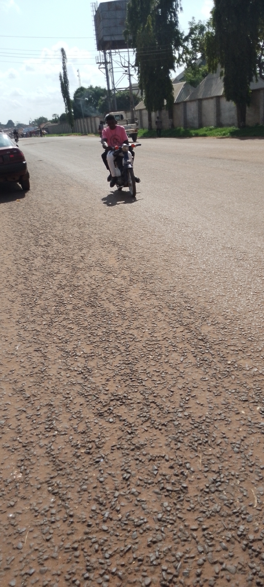 A person wearing a pink shirt and white pants is riding a motorcycle on a road. The motorcycle is dark colored. The person is looking forward. The road is paved with small stones. In the background, there is a tall water tower, trees, and buildings. There is also a dark colored car on the left side of the road. The sky is cloudy. The scene appears to be in a town or city in Adikpo, Nigeria. The time of day is daytime. The weather appears to be clear and sunny.