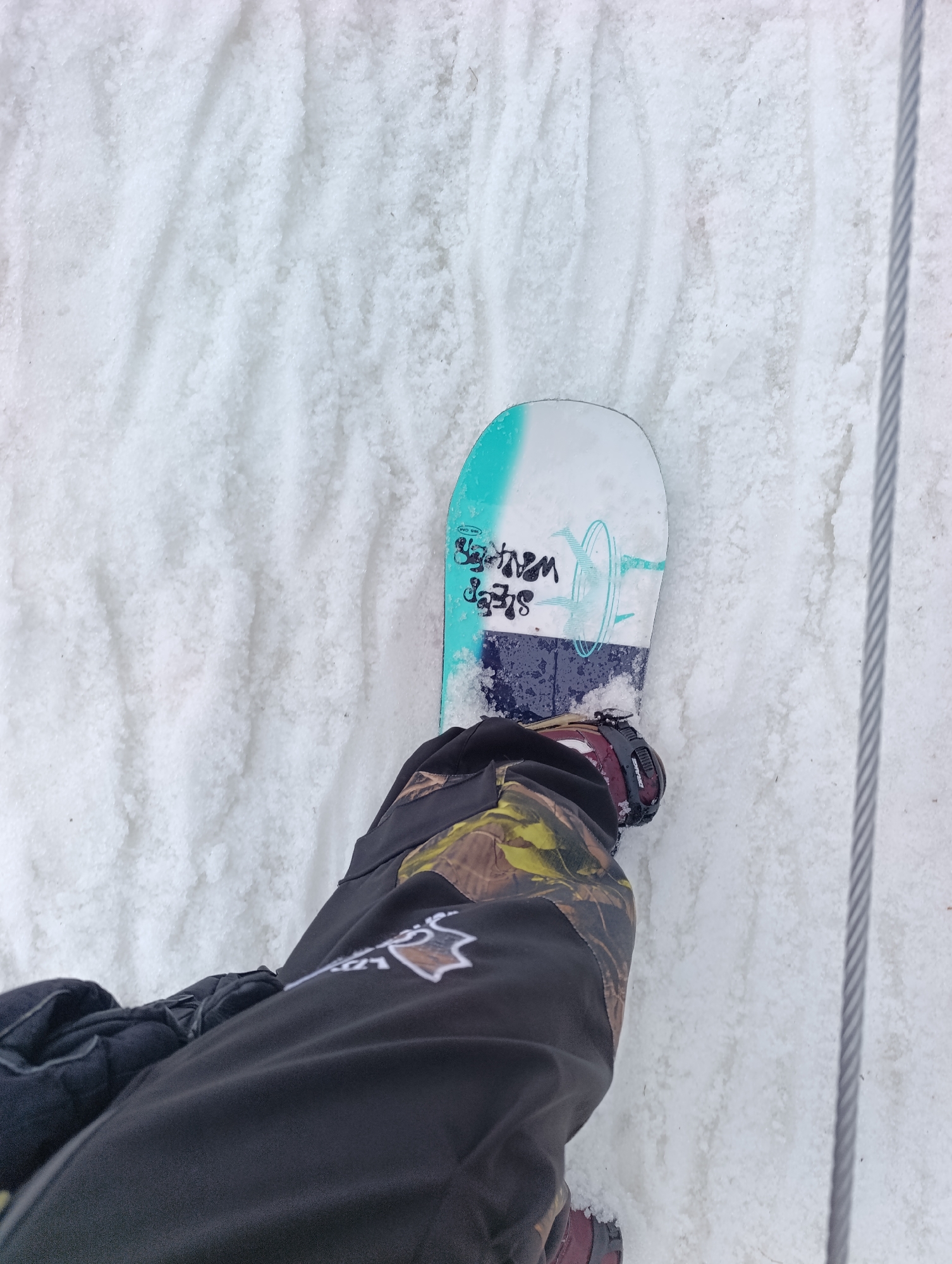 A top-down shot shows a person standing on a snowboard in a snowy environment. The snowboard is predominantly white and has some writing on it in black and turquoise. The person is wearing snow pants that are black with brown and yellow camouflage patterns. A cable runs vertically along the right side of the image. The snow appears to be packed and slightly textured. The exact location within Krasnoyarsk, Russia is not discernible from the image alone.