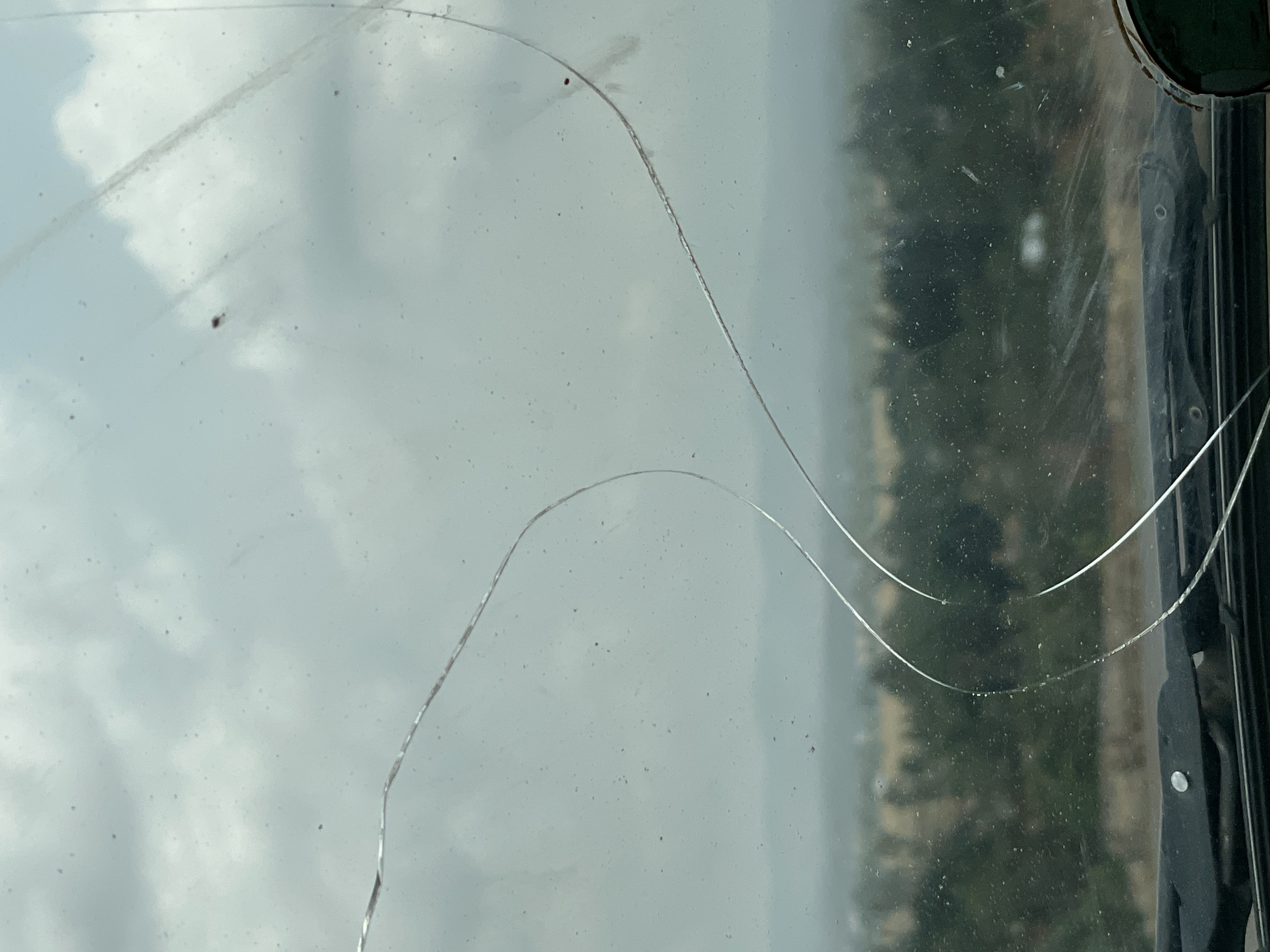 The image shows a view through a cracked windshield. The windshield is dirty with numerous small specks and two prominent cracks. The view through the windshield includes a hazy or cloudy sky in the upper portion and a blurred landscape with trees or vegetation in the middle to lower portion. The vehicle's frame is visible on the right side of the image. The city and specific location in Vom, Nigeria is not evident in the image.