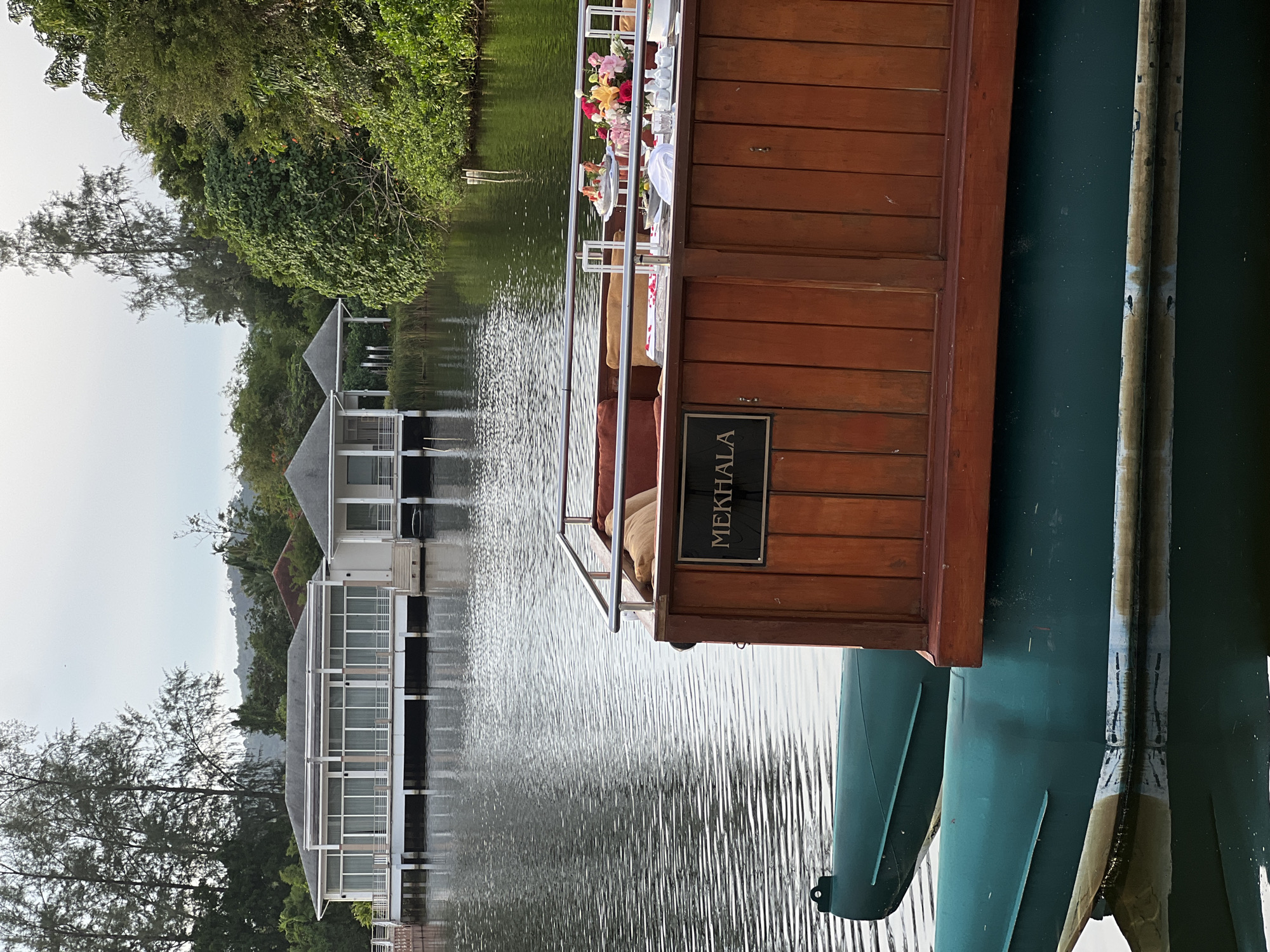 An outdoor shot captures a boat named "MEKHALA" on a calm body of water. The boat has a wooden superstructure and two green pontoons visible below the waterline. The superstructure is decorated with cushions and a table set for a meal, complete with flowers and food. In the background, several modern white buildings with large windows are situated along the shore, surrounded by lush green trees and foliage. The sky above is overcast. The scene appears to be a serene waterfront setting, possibly a resort or private property.