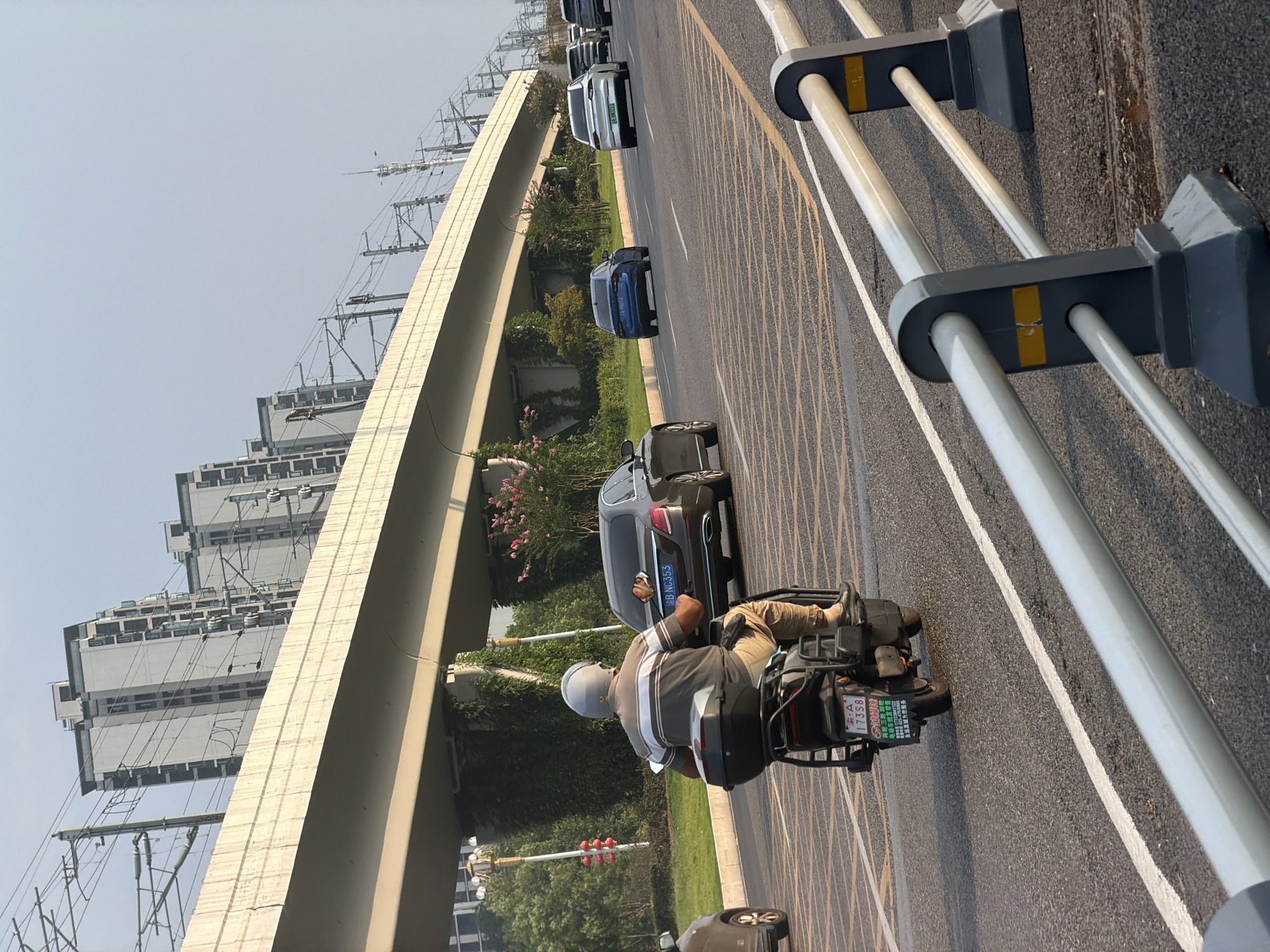 The image shows a street scene in Jiangjin, China, with a multi-level overpass dominating the background. Apartment buildings are visible beyond the overpass. A man is riding a scooter in the foreground, wearing a helmet and a striped shirt, with a black storage box on the back of the scooter. Several cars are on the road, including a black sedan with a license plate that reads "38-NC353," a blue sedan, and a white sedan. The road has yellow and white lane markings. A metal guardrail is visible in the lower right corner of the image. The lighting suggests it is daytime, likely late afternoon given the long shadows.