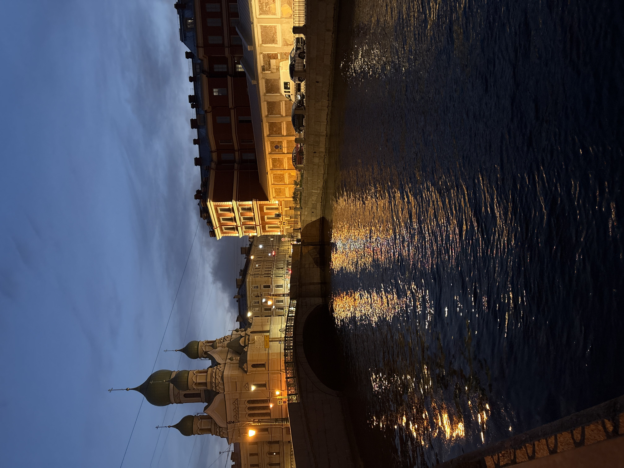 The image shows a nocturnal cityscape in Saint Petersburg, Russia. A river dominates the right side of the frame, reflecting the illuminated buildings along its bank. The buildings are lit with a warm glow, contrasting with the dark water. On the left, a Russian Orthodox church with distinctive onion domes is visible. The sky is a twilight blue, adding to the ambiance. A bridge crosses the river in the center of the frame, with the reflection of the cityscape creating symmetry. Cars are parked along the riverbank.