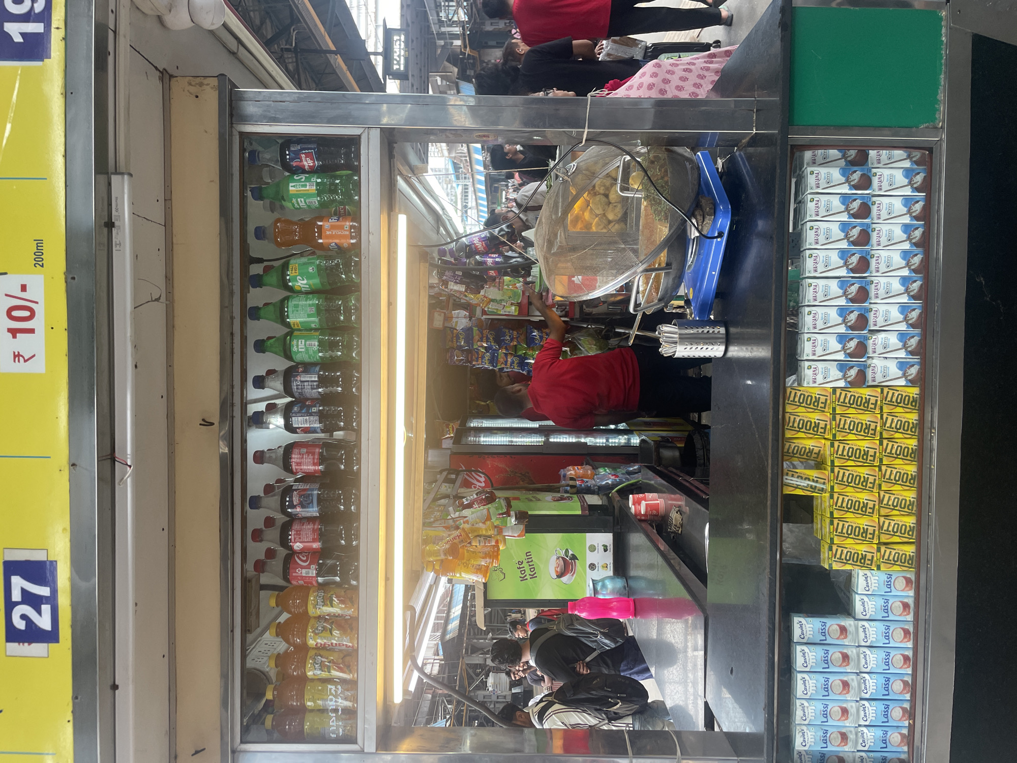 This image captures a street-side food and beverage stall, likely located in Mumbai, India, given the context clues of the Hindi signage and the typical appearance of such stalls in Indian cities. The stall is positioned outdoors, possibly within a bustling market or a train station, indicated by the elevated railway track structure visible in the background.
In the foreground, a long, dark countertop displays various items for sale. At the bottom of the stall, an illuminated glass-fronted refrigerator showcases rows of packaged drinks, including cartons labeled "Lassi," "FROOTI," and "CHAAS." To the left, a bright pink plastic water bottle rests on the counter, next to a blue container.
Behind the counter, a vendor in a red shirt is actively serving customers, with his hand reaching towards a tray of fried snacks covered by a mesh dome. A few customers, including a woman in a pink patterned dress and others carrying bags, are visible, some looking towards the stall. In the mid-ground, a green refrigerator or display unit features the prominent text "Kafe Kartin" and an image of tea, suggesting coffee and tea are also available.
Above the counter, shelves are stocked with a variety of bottled beverages, including soft drinks like Coca-Cola, Sprite, and Thums Up, along with other colorful liquid products. Bright yellow signage with the number "27" and pricing information like "₹ 10/-" further identifies the stall and its offerings. The overall scene depicts a vibrant and busy point of sale, typical of urban India.