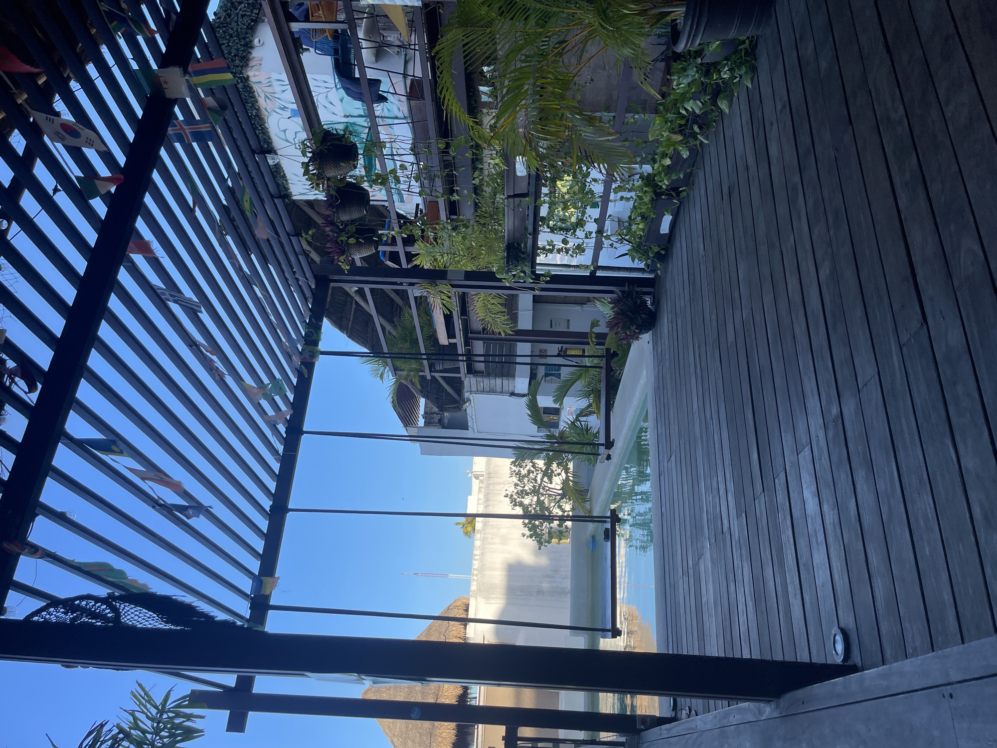 The image shows an outdoor rooftop scene in Playa del Carmen, Mexico. A wooden deck extends across the right side of the frame, with a pool visible beyond a glass barrier. Above the deck is a pergola structure decorated with hanging plants and small international flags. Reflections on the glass show nearby buildings and the bright blue sky. The image is taken from an elevated position.