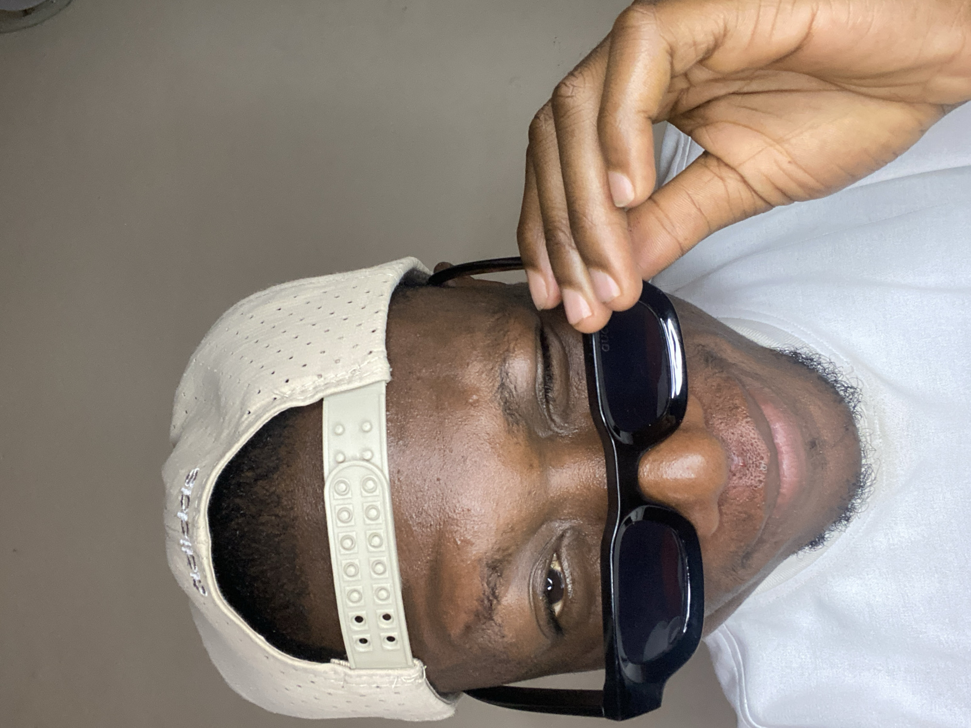 A man with dark skin wears sunglasses and a white perforated baseball cap. He is touching the sunglasses with his right hand. He is wearing a white t-shirt. The background is a light beige wall. The location of the image is undetermined.
