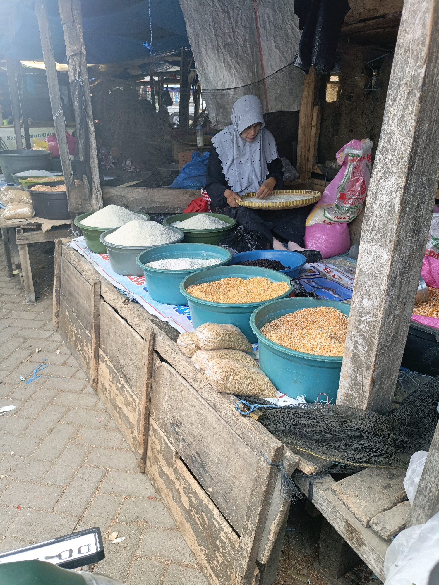 The image depicts an outdoor market stall in Bulukumba, Indonesia, characterized by rustic wooden structures and a blue tarp roof. A woman, wearing a light blue hijab and dark clothing, is seated behind a collection of large turquoise and green plastic tubs filled with various grains, including what appears to be rice and corn. She is holding a woven tray, also filled with grain, in her lap. Several sacks of grain are visible, along with a pink plastic bag and a blue plastic bag. The ground is paved with interlocking bricks. The stall appears to be part of a larger market, with other stalls and structures visible in the background. The overall impression is one of a functional, everyday market scene.