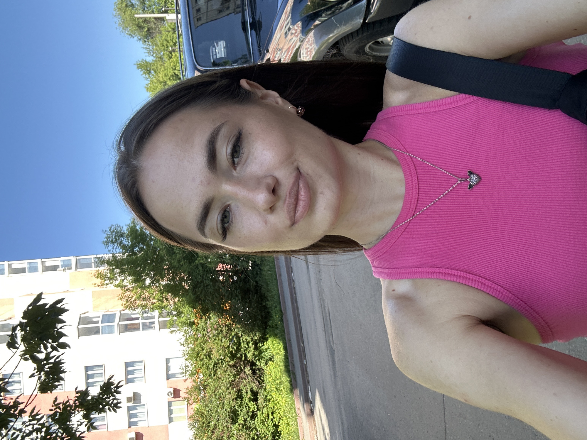 An outdoor, eye-level medium shot shows a young woman from the chest up, posing for a selfie in an urban environment. The woman has long, dark brown hair and is wearing a bright pink tank top and a silver necklace with a winged heart pendant. She has a subtle smile and is looking directly at the camera. Behind her, a black SUV is parked on the street, and a modern apartment building with light-colored walls and numerous windows is visible. Lush green bushes and trees line the sidewalk in front of the building, and the sky is a clear, bright blue. The overall scene suggests a sunny day in a residential area.