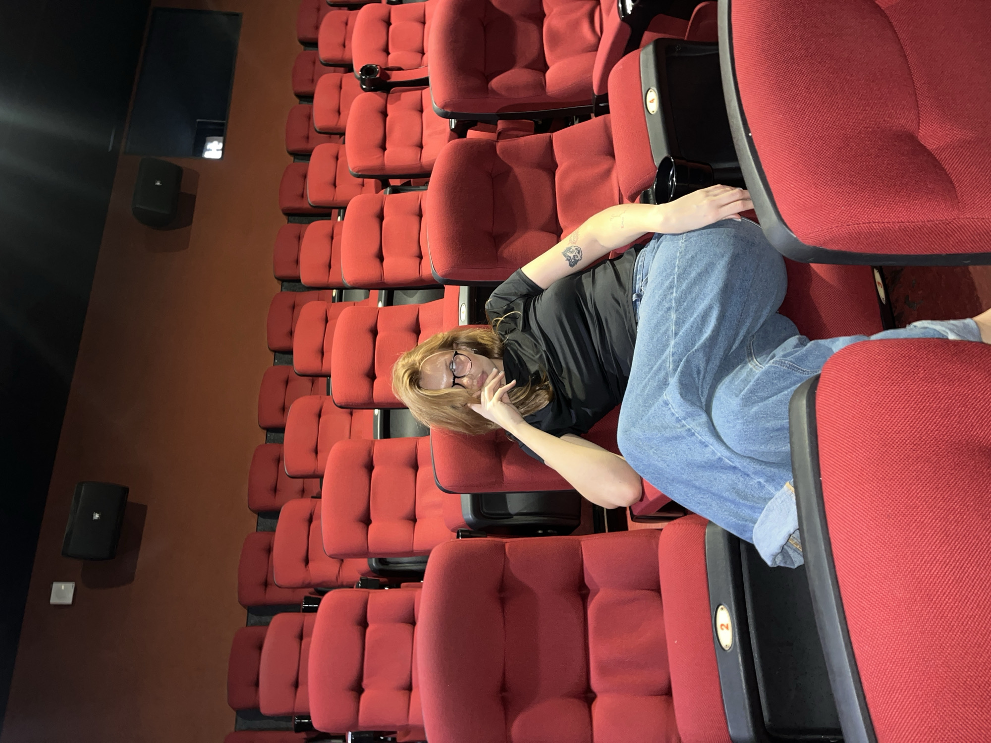 A young woman with long, reddish-brown hair and glasses is seated in an empty movie theater. She is wearing a black t-shirt and loose-fitting blue jeans. Her left arm rests on her bent leg, and her hand is near her face. A tattoo is visible on her left forearm. The seats in the theater are bright red, and the walls are a dark reddish-brown. Speakers are mounted on the wall above the seats. The image appears to be taken from a slightly elevated angle, looking down at the woman. The overall atmosphere is calm and quiet, suggesting an unoccupied theater. This scene is located in the city of Chaikovsky, Russia.