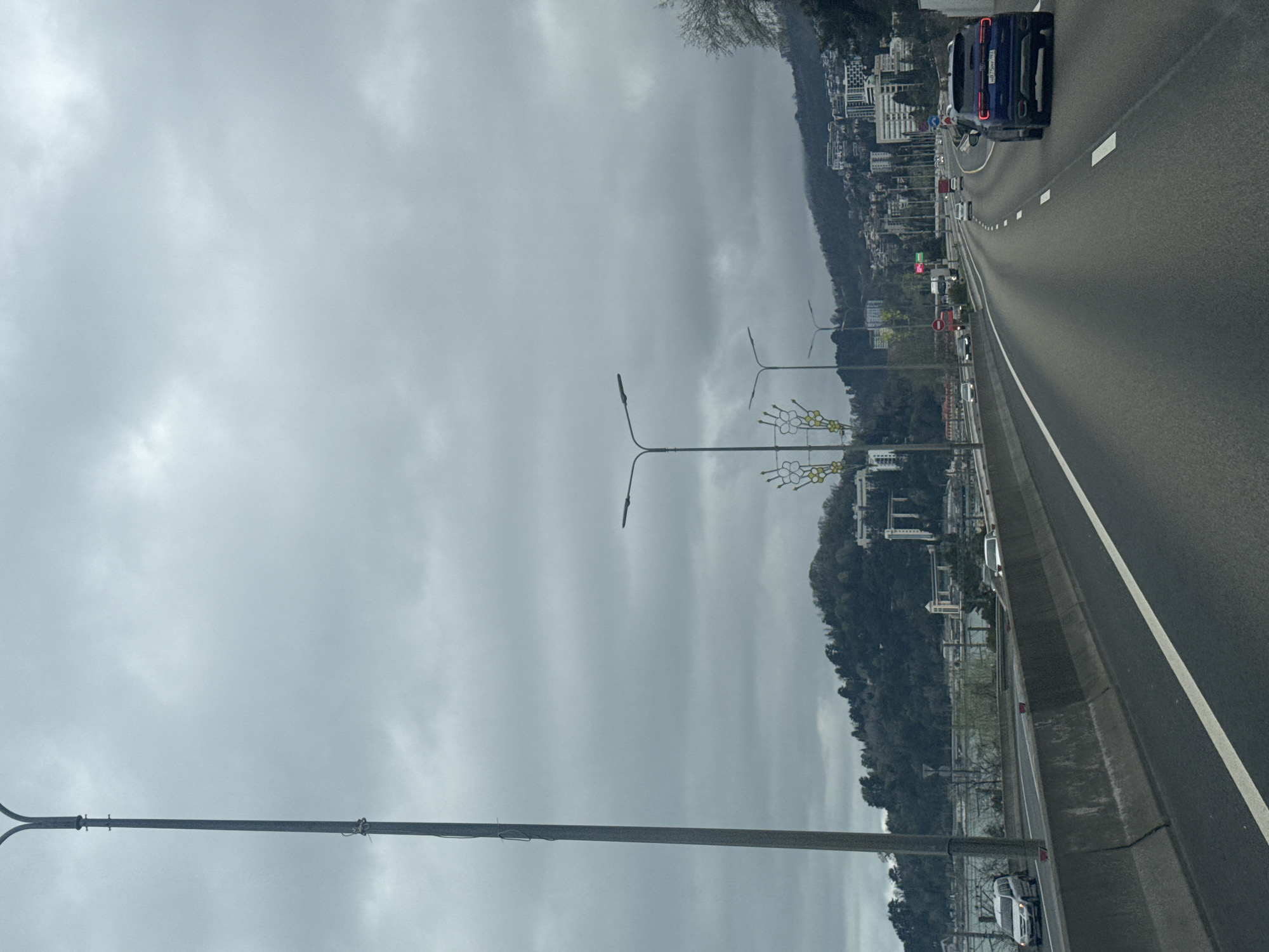 The image shows a highway in Sochi, Russia, under a cloudy sky. A blue car is traveling on the right side of the road. Several other cars are visible further down the road. On the left side of the highway, there are trees, buildings, and streetlights. In the far distance, a mountain can be seen.