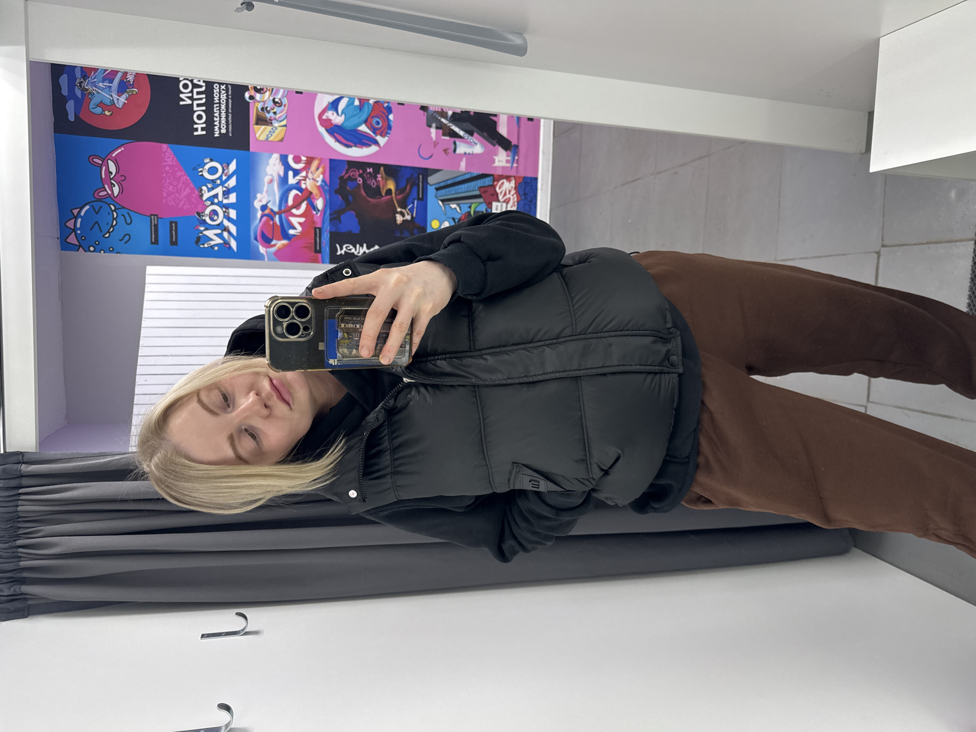 The image shows a woman taking a selfie in what appears to be a dressing room. She is fair-skinned with blonde hair and is wearing a black vest over a black hoodie and brown sweatpants. She is holding a cell phone in her left hand to take the photo. The dressing room has a gray curtain, white walls, and what appears to be a tiled floor. There are colorful cartoon-like posters on the wall behind her. The posters have text in Cyrillic, suggesting the location is likely in a Russian-speaking area. Based on this and the prompt, the location is presumed to be in Saint Petersburg, Russia, although there are no specific landmarks to confirm this.