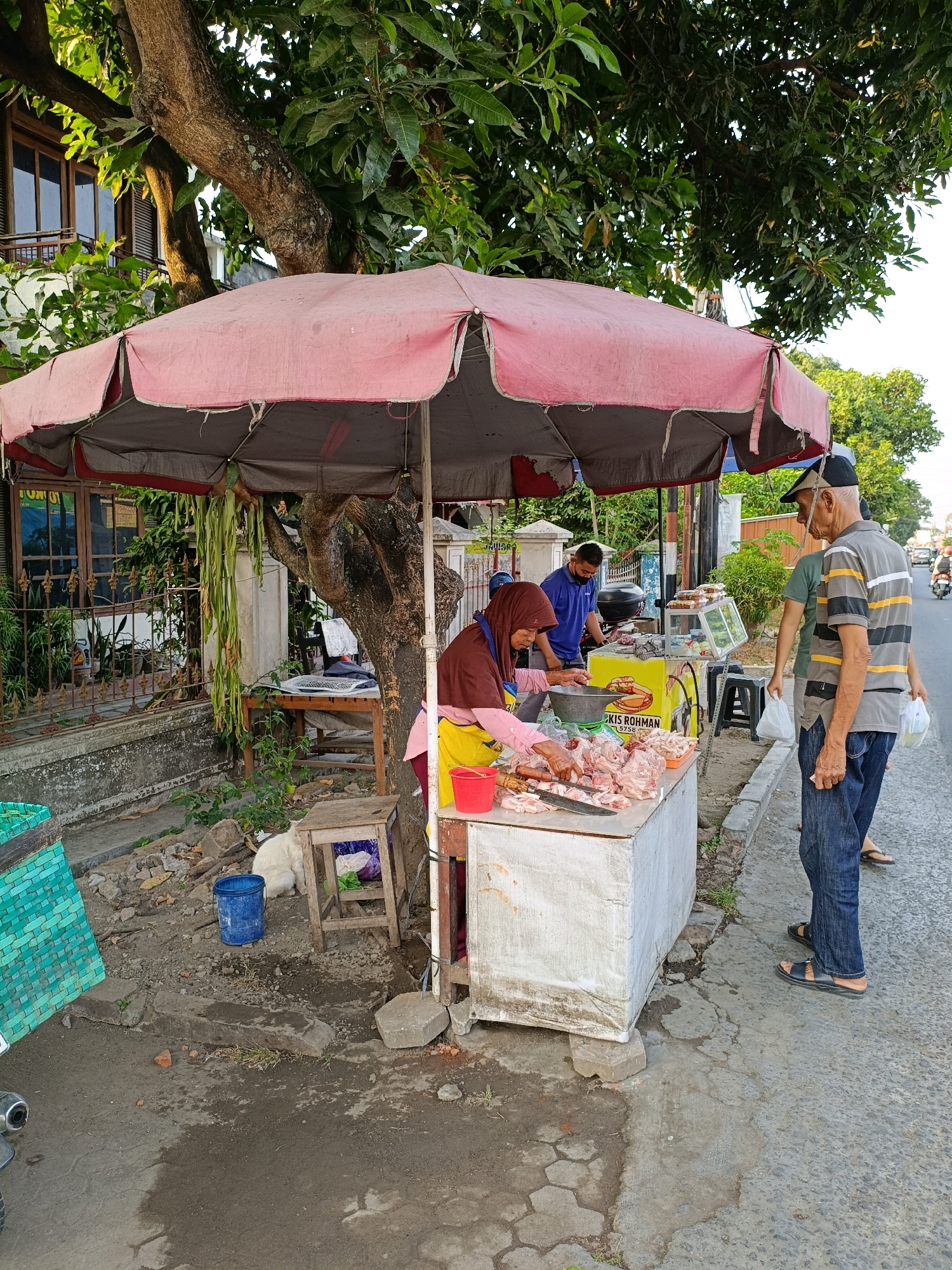 The image depicts an outdoor meat stall under a large, pink and grey umbrella, likely situated on a roadside in Purwomartani, Indonesia. A woman, wearing a dark hijab and a pink apron over a yellow garment, is actively cutting raw chicken pieces on a white stall. In front of her, a large quantity of prepared chicken is displayed on the stall's surface. To her right, an elderly man, wearing a black cap, a striped shirt, and blue jeans, is standing and facing the stall, appearing to be a customer. Behind him, another man in a green shirt is also present, holding a white plastic bag. Further to the left, a man in a blue shirt is visible near a motorcycle, which is parked behind the stall. The stall itself is a white structure, possibly a cart or table, with a yellow side panel featuring some text. Various items are scattered around the immediate area, including a blue bucket, a small wooden stool, discarded bricks, and a green woven basket to the left. A large tree with a thick trunk shades a portion of the scene, with its branches and leaves extending overhead. In the background, a building with wooden-framed windows and a fence is visible, along with other trees and a road with passing vehicles. The ground is a mix of asphalt and dirt, with some puddles and debris.