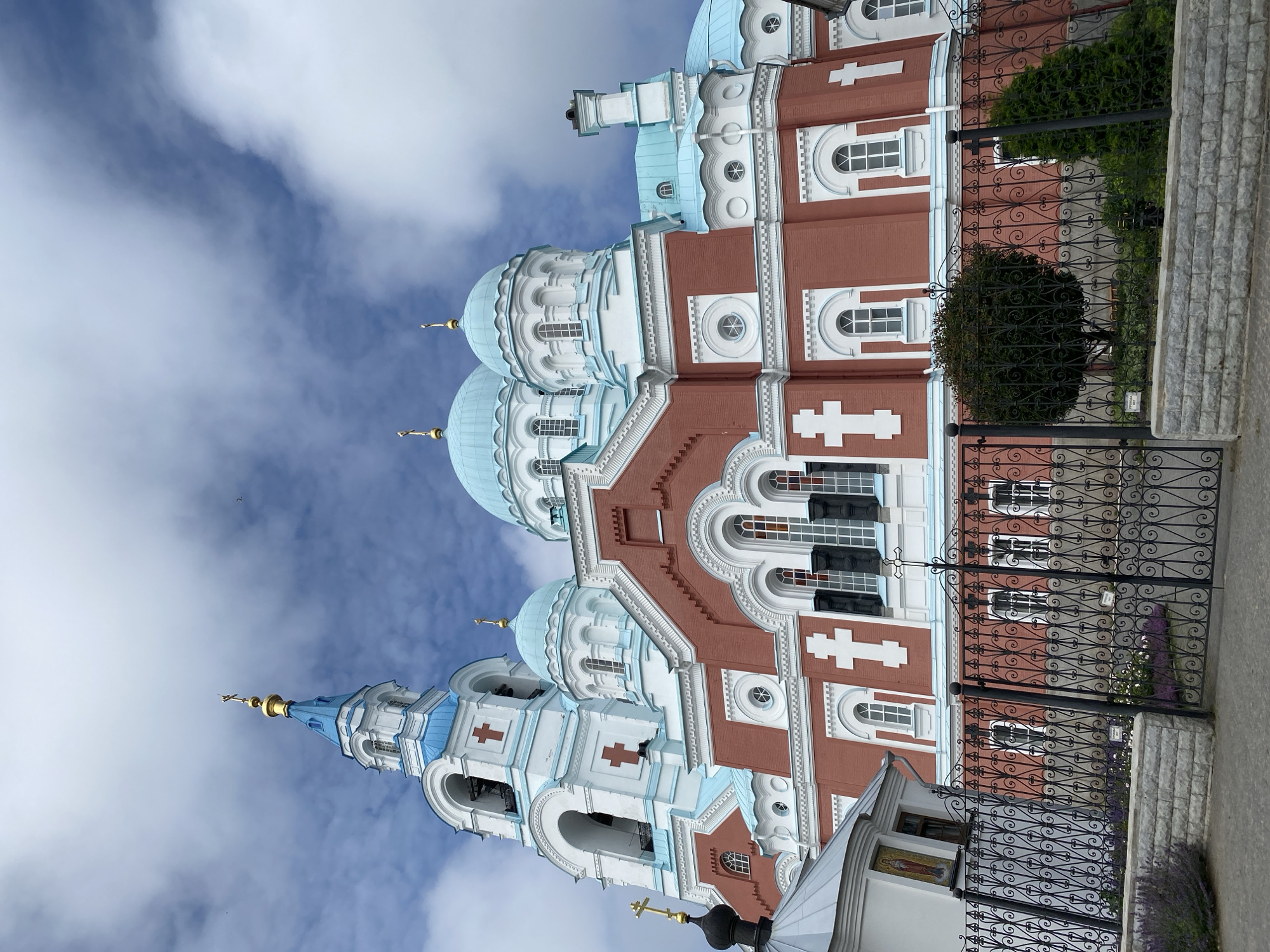 The image shows a large, red and white Orthodox church with blue domes, located in Sortavala, Russia. The church has a tall bell tower with a blue spire topped with a golden cross. The main building has several domes, also blue, with golden crosses. White decorative elements and red crosses adorn the red facade of the church. A black wrought-iron fence is in the foreground, with a stone wall and some greenery behind it. The sky is partly cloudy with a blue hue.