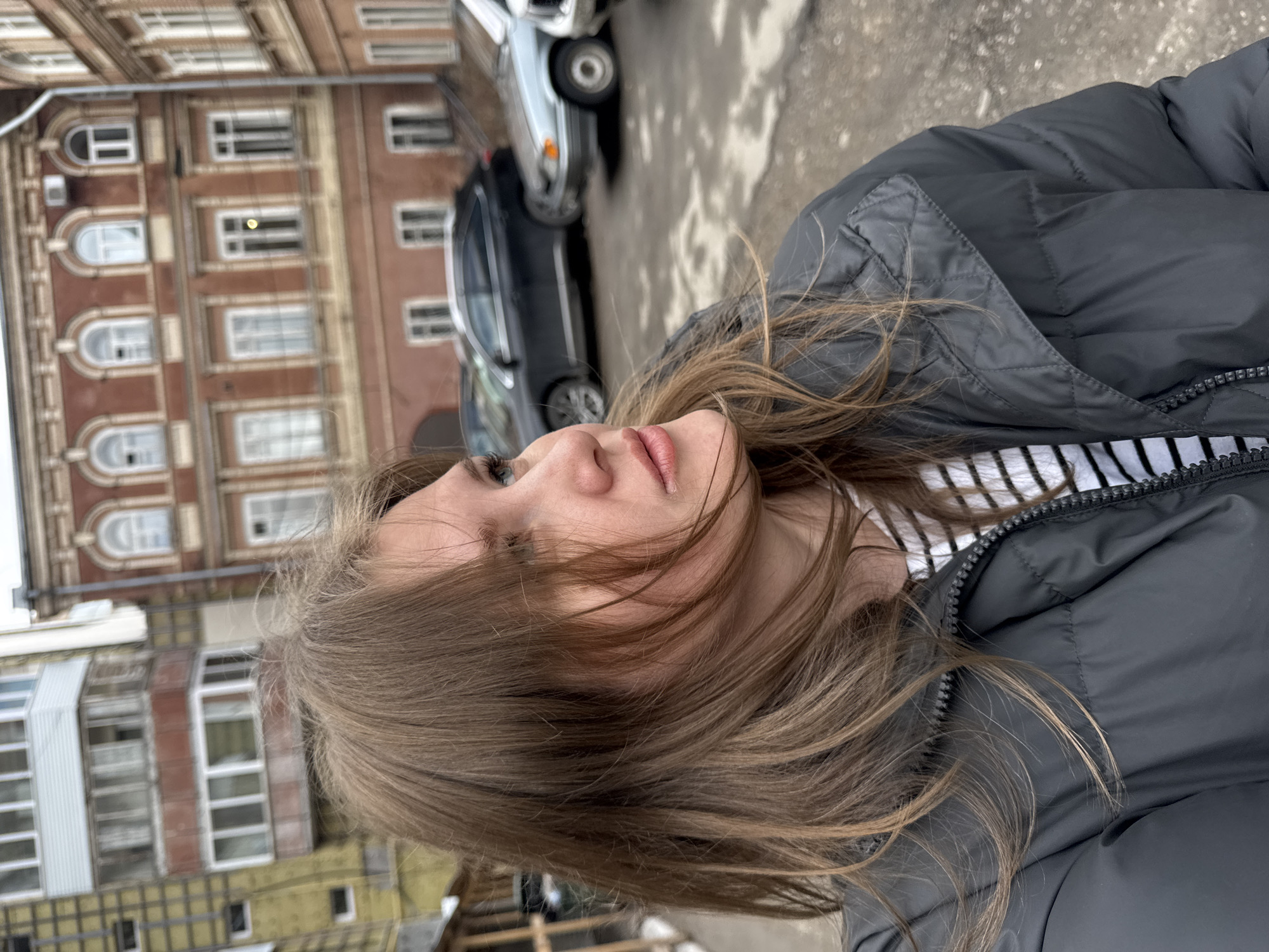 A woman with brown hair, fair skin, and neutral makeup stands in an urban setting. She is wearing a black puffer jacket over a black and white striped shirt. The background shows a street with a brown brick building with multiple windows and a yellow building, alongside a few parked cars. The photo was taken in the city of Perm, Russia.