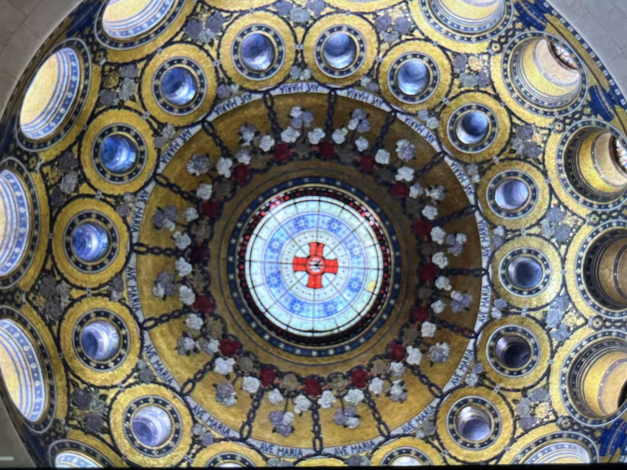 The image shows the ornate ceiling of a religious building, likely a church or cathedral. The ceiling has a circular design with multiple concentric rings. The outermost rings feature circular medallions with blue and gold accents. Closer to the center, there are embellishments with floral designs in red, white, and gray. The words "AVE MARIA" are inscribed along parts of the design. The very center of the ceiling features a circular stained-glass window with a red cross in the middle. While the image is provided in the context of Tinaquillo, Venezuela, the information available does not specify the exact location within the city.