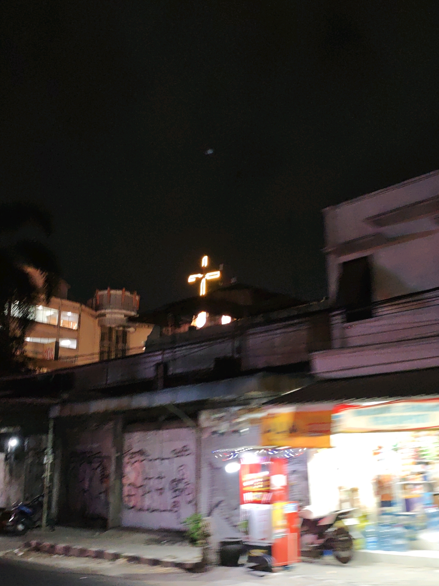 This image captures a nighttime street scene in Kediri City, Indonesia. A prominent, illuminated cross glows brightly against the dark sky, positioned above a building structure. To the left, a multi-story building with lit windows is visible, partially obscured by dark foliage. In the foreground, a row of shops lines the street. One shop features a colorful sign and a display of goods, including bottled water. A motorcycle is parked near the shops, and graffiti adorns the walls of a nearby structure. The overall impression is that of an urban environment at night, with a mix of religious symbolism and commercial activity.