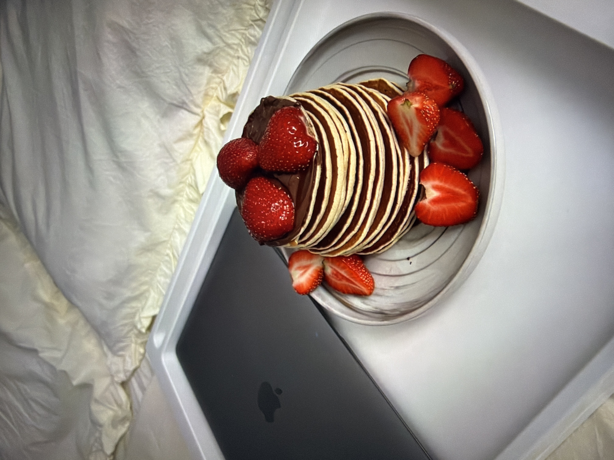 A stack of chocolate-covered pancakes topped with strawberries and surrounded by sliced strawberries sits on a grey and white patterned plate. The plate is on a white tray, next to a closed dark grey laptop with a glowing apple logo. The tray is positioned on a bed with rumpled cream-colored sheets and a pillow. The scene appears to be indoors, likely a bedroom. There is no information to suggest a location in Kyiv, Ukraine.