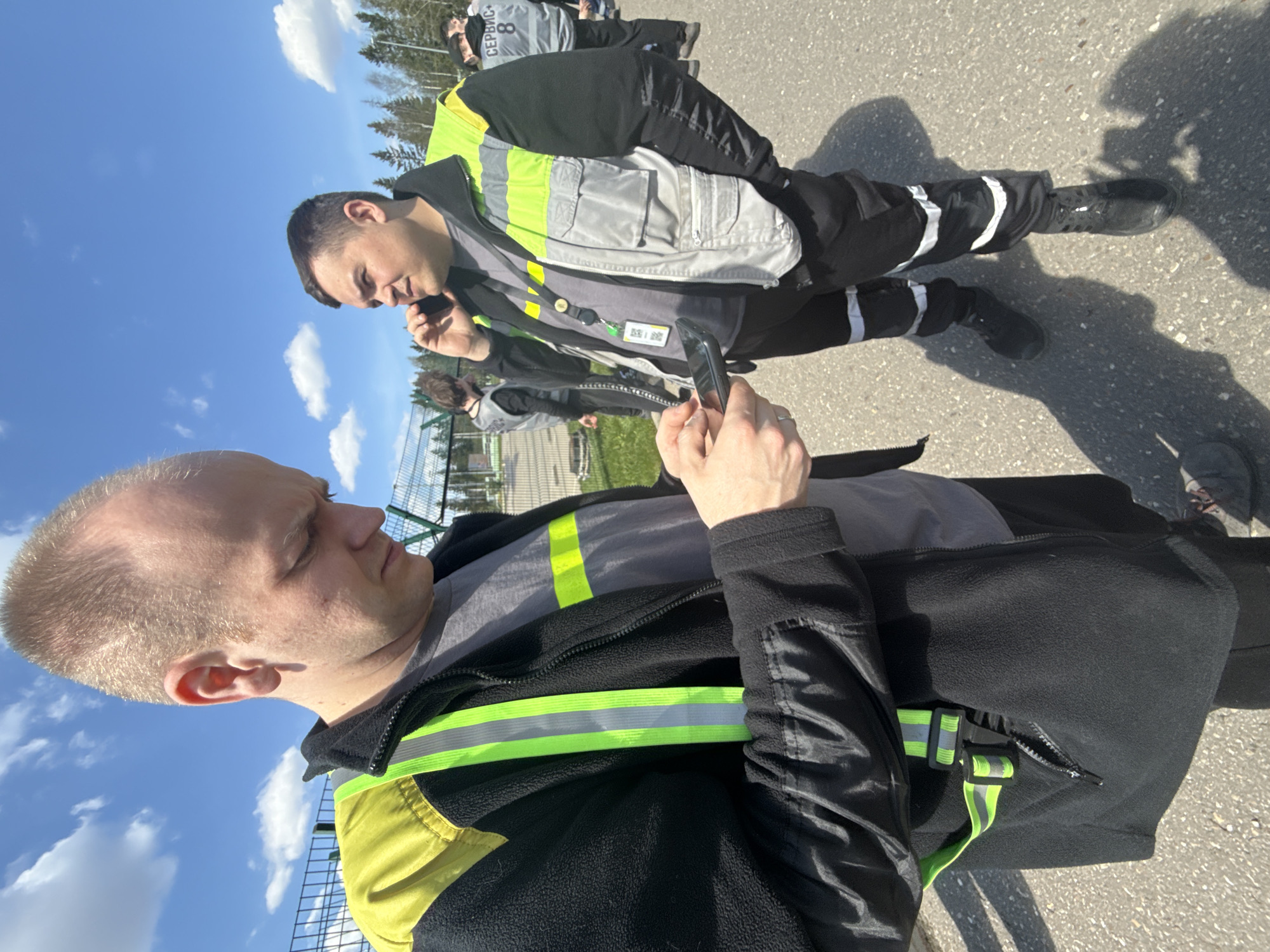 The image shows two men standing on a paved surface. Both men are wearing clothing with reflective high-visibility stripes. One man is holding a mobile phone, possibly looking at it. The other man appears to be speaking on a phone. The location is presumed to be Lupanovo, Russia. The background includes a fence, trees, and a partly cloudy sky. Other people are present in the background.