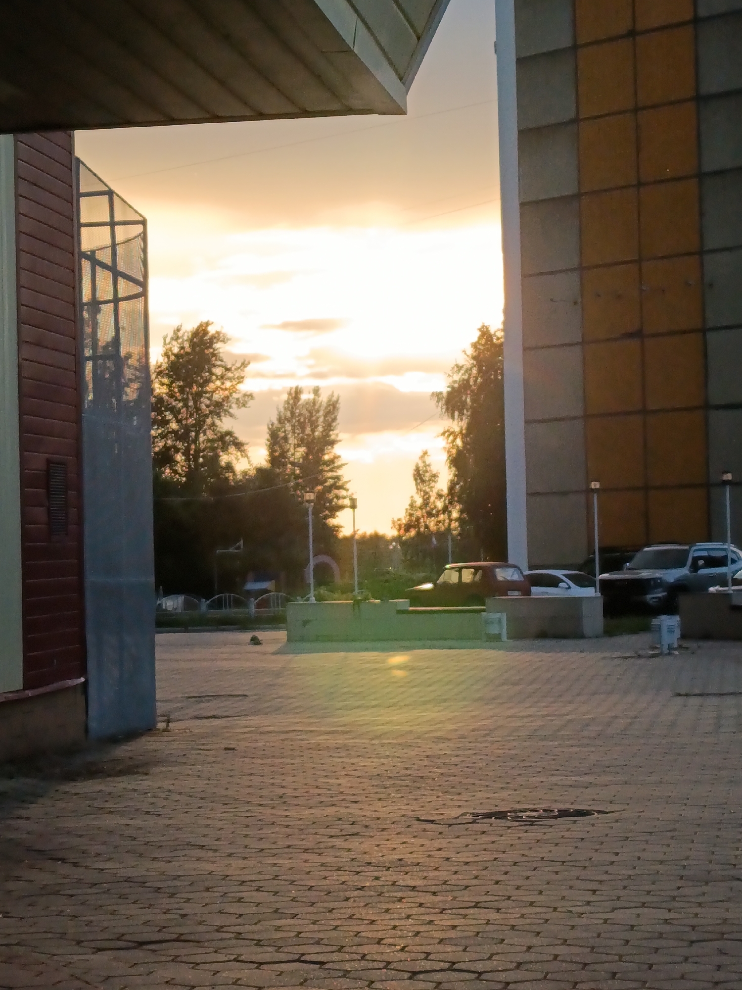 The image depicts an outdoor scene at dusk, possibly in the city of Chaikovsky, Russia, characterized by paved ground with cobblestone-like patterns. On the left, a building with red siding and a grey, ribbed architectural element is partially visible. To the right, a multi-story building with a facade of alternating yellow and grey square tiles dominates the background. A gap between these two structures reveals trees and the sky. The sky is illuminated by a sunset, with warm orange and yellow hues and scattered clouds. Several cars are parked in the area visible through the gap, including a dark red hatchback, a white sedan, and a dark SUV. Streetlights are also present. The foreground pavement shows a manhole cover. The lighting suggests the sun is low on the horizon, casting long shadows and creating a warm, diffuse light across the scene.