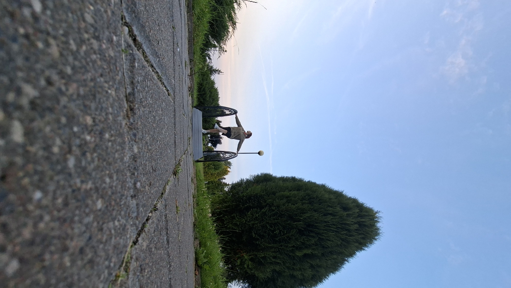 A low-angle, wide shot captures a person standing on a decorative metal bridge in Myadzyel, Belarus. The person, dressed in a green jacket and dark shorts, is balanced on one leg with arms extended, seemingly posing. The bridge spans a paved pathway made of interlocking gray stones, which fill the foreground. Behind the bridge, lush green grass and manicured bushes line the path, leading towards a very large, cone-shaped evergreen tree. The sky above is a clear, pale blue with a few wispy white clouds. The lighting suggests it is late afternoon or early evening.