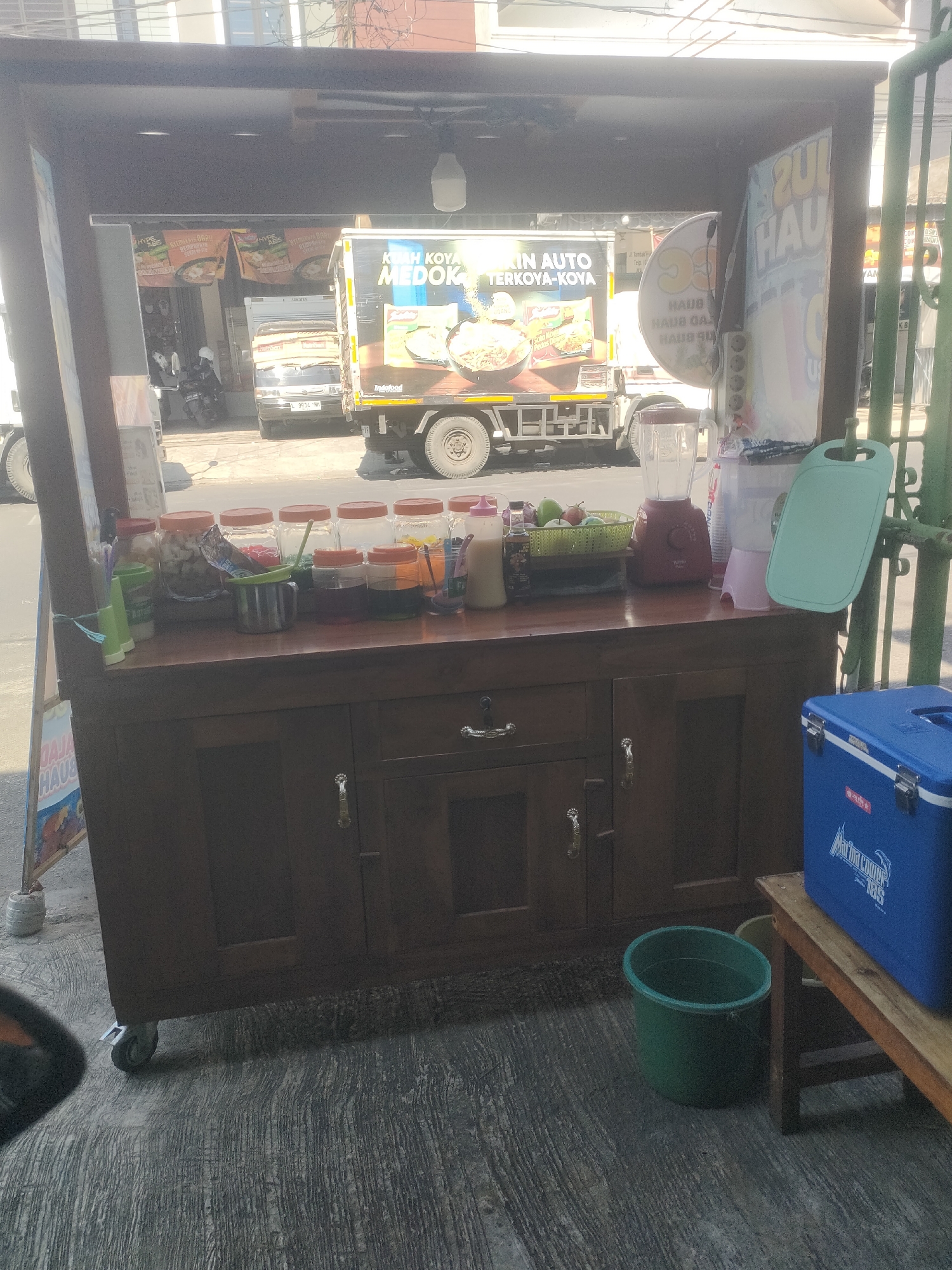 This image depicts a food cart situated on a sidewalk in Surabaya, Indonesia. The cart is made of dark wood and has a countertop displaying various items, including jars of ingredients, a blender, and cups. Behind the cart, a delivery truck with "Indomie" branding is visible. The environment suggests a street setting with buildings and a road in the background. The cart is equipped with wheels, indicating mobility.
