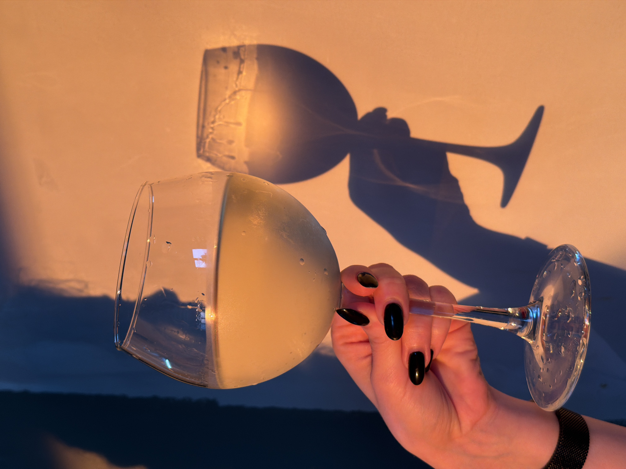 The image captures a close-up, artistic shot of a person's hand holding a large wine glass filled with a light-colored beverage, likely a cocktail or white wine. The hand, adorned with glossy black nail polish and a dark mesh watch or bracelet on the wrist, holds the clear stem of the glass. The liquid inside the glass appears somewhat opaque and frothy on the surface, with small water droplets visible on the exterior of the glass.
The scene is dramatically lit, suggesting either golden hour during sunset or sunrise. Warm, orange-yellow light bathes the upper right portion of the background, which appears to be a wall or flat surface. A prominent, dark shadow of the wine glass and the hand holding it is cast sharply across this warm background, displaying detailed contours of the glass and fingers. The interaction of light and shadow is a key element of the composition.
In contrast to the warm, illuminated background, the lower left and bottom portions of the image feature a darker, cooler-toned background, transitioning from deep blue to almost black. This suggests a view outwards, possibly a twilight sky or landscape seen through a window, or simply a deep shadow area within the room.
The overall mood is contemplative and serene, emphasizing the interplay of light, liquid, and form. No text is visible in the image. While the location context of Chaikovsky, Russia is provided, there are no visual cues within the image itself to confirm this specific geographical location.