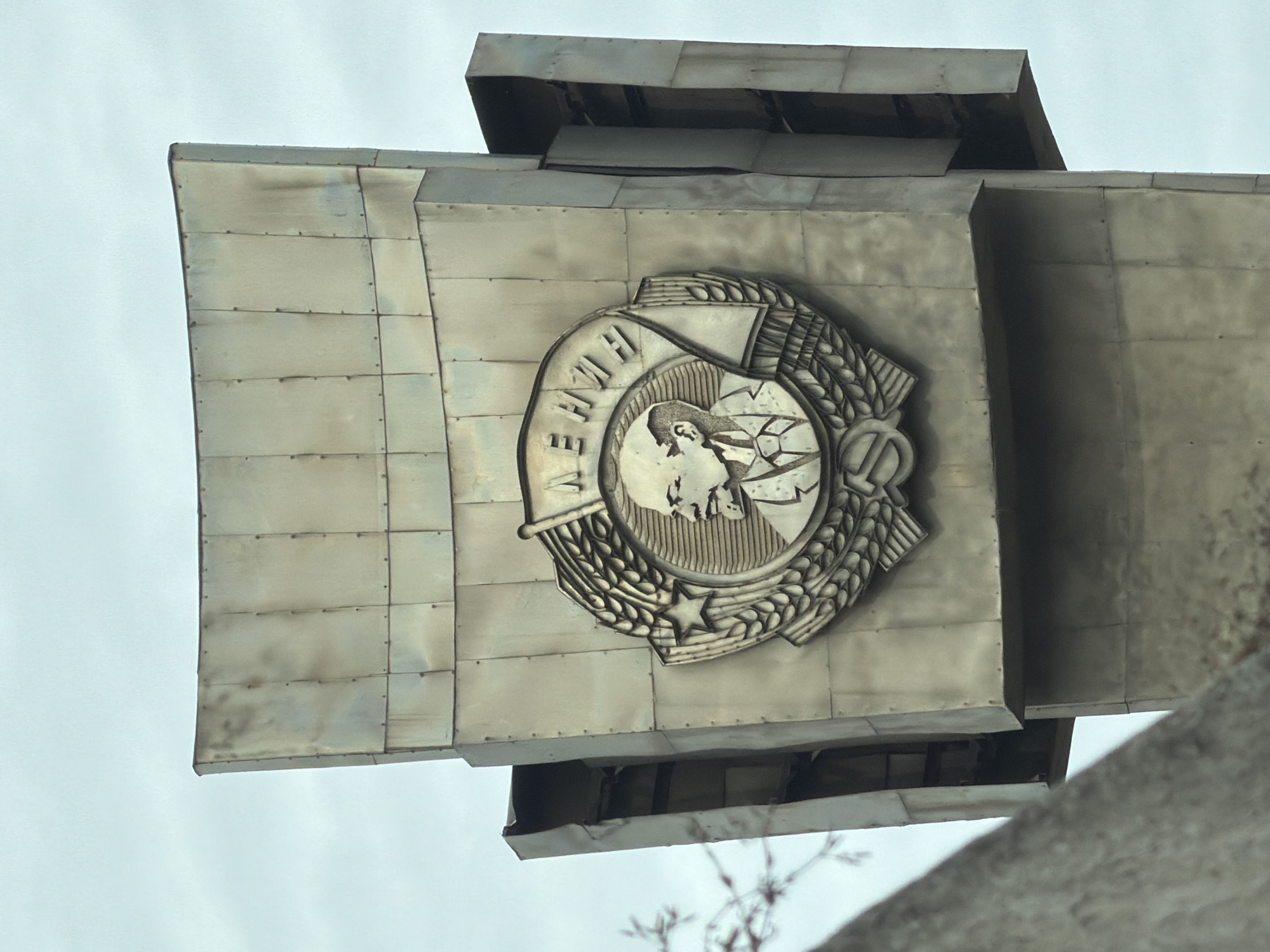 The image depicts a close-up, low-angle view of a monument in Perm, Russia. The monument features a large, metallic emblem of the Soviet Union prominently displayed on a block-like structure composed of rectangular panels. The emblem includes a profile of Vladimir Lenin, a globe, a hammer and sickle, and ears of wheat. The sky is overcast and pale. In the foreground, out-of-focus architectural elements and bare tree branches can be seen.