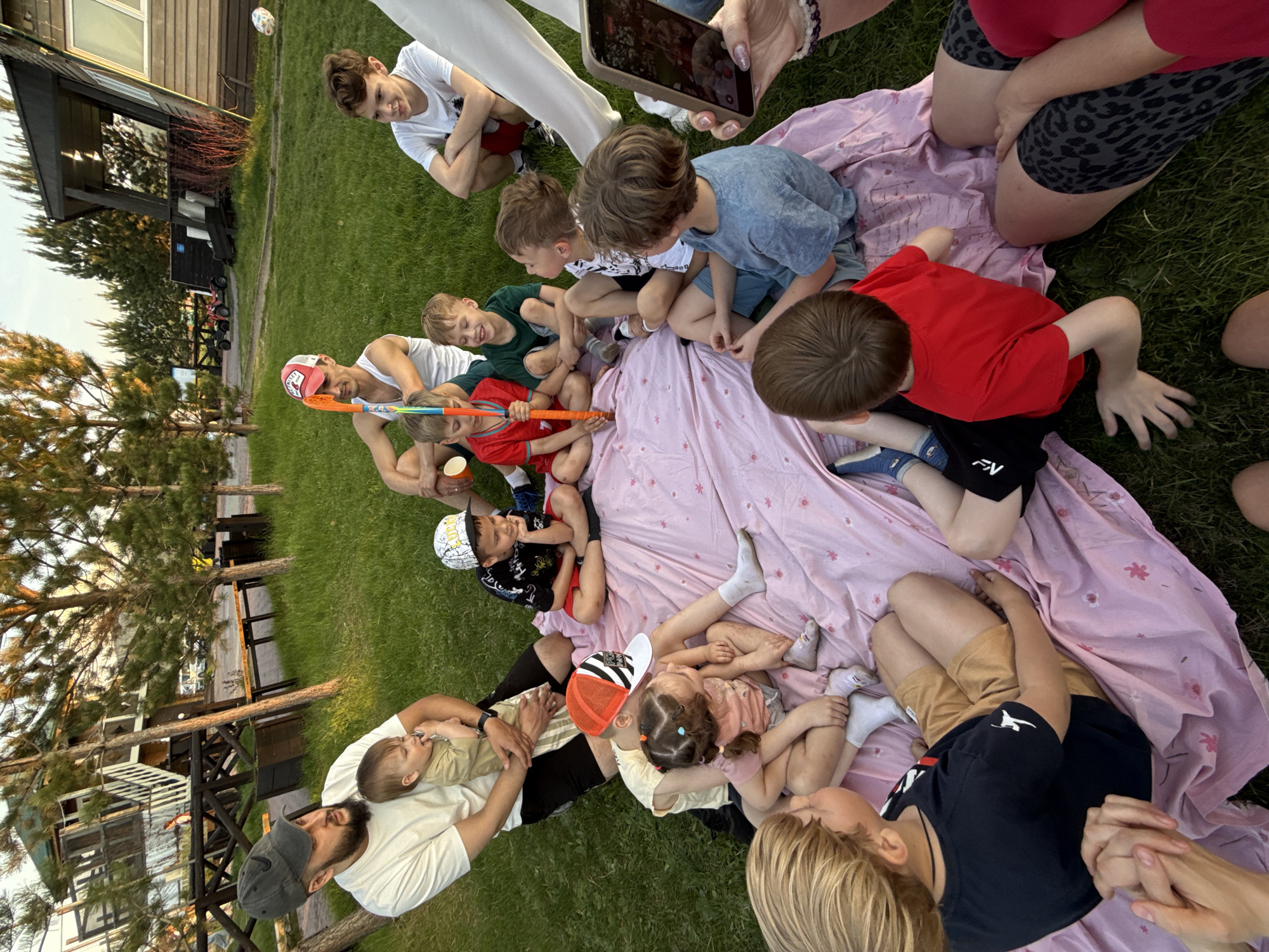 The image shows a group of people on a lawn in what appears to be a residential area. Based on the prompt, the location is Кузнецово, Russia. A group of children and adults are gathered on and around a pink blanket with floral patterns. Some of the adults are holding or interacting with the children. A man is holding a baby. One adult is holding a phone, possibly taking a picture. One man is holding a colorful stick. The environment includes green grass, trees, and buildings in the background.