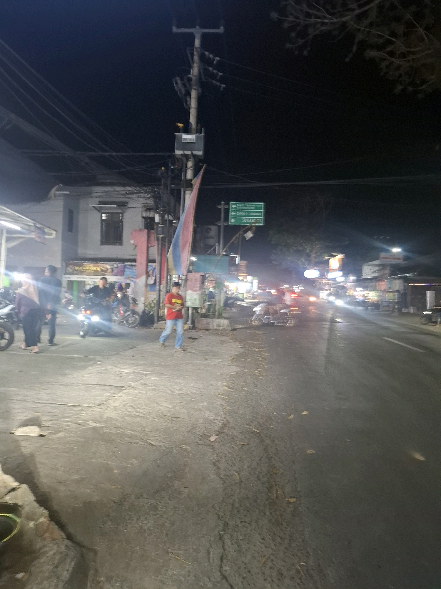 The image shows a street scene at night in Karangtengah, Indonesia. A busy street is lined with buildings, some of which appear to be shops or food stalls, illuminated by various lights. Several people are visible on the sidewalk and street, including a person in a red shirt walking on the sidewalk and a group of individuals near motorcycles. Motorcycles are a prominent mode of transportation, with several parked and others being ridden. Traffic, including cars and motorcycles, is present on the street, with headlights and taillights indicating movement. Overhead power lines and a utility pole are also visible. A green directional sign is mounted on a pole, indicating directions to different locations. The overall impression is of a vibrant urban environment at night, with a moderate level of activity.
