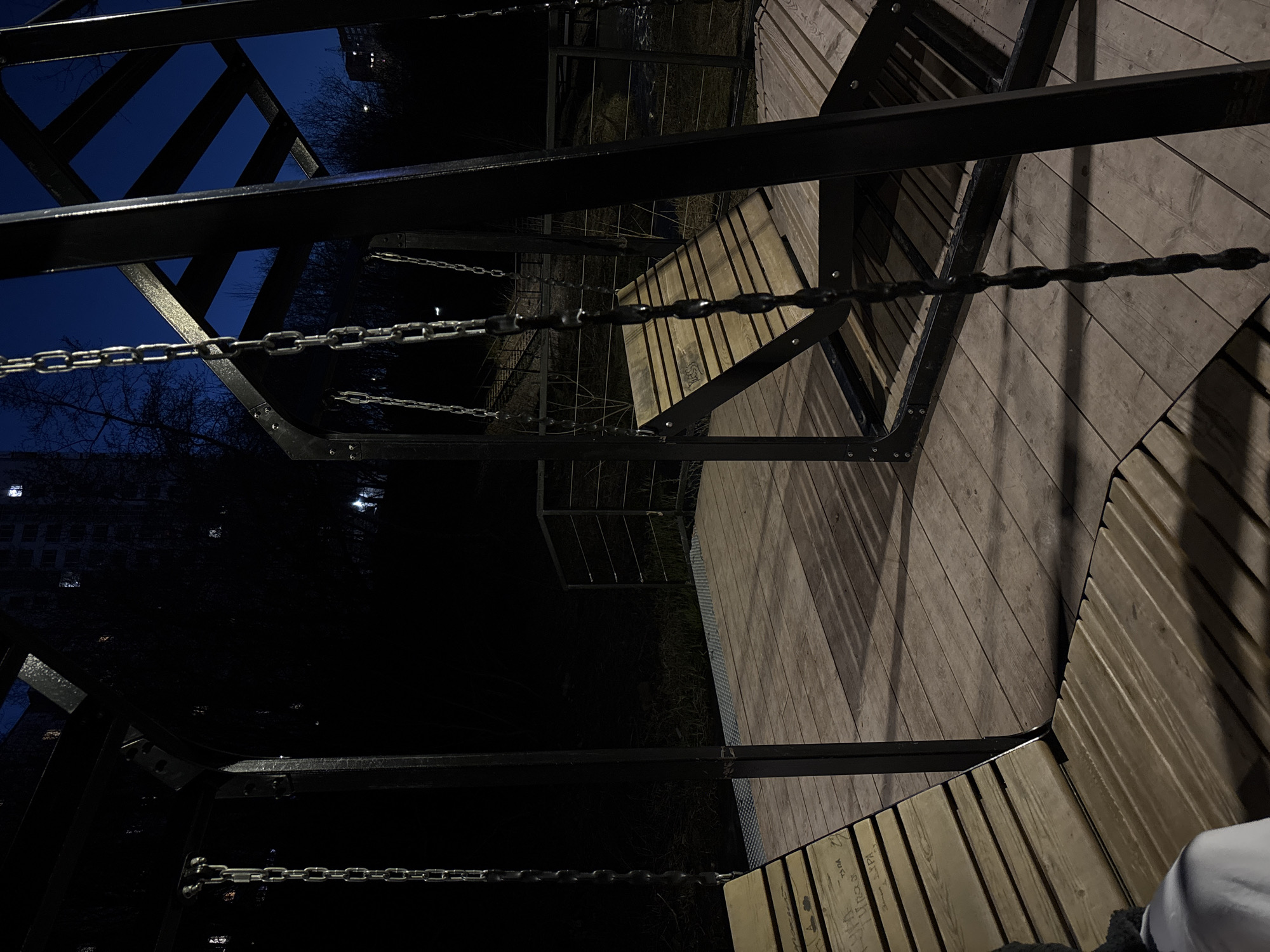This image captures a nocturnal scene of an outdoor public space in Perm, Russia. The primary focus is a modern architectural structure made of dark metal frameworks and light-colored wooden surfaces.
The structure features angular dark metal beams and supports that crisscross the frame, creating dynamic lines. Several robust metal chains are prominently displayed, stretching horizontally, likely serving as barriers, handrails, or decorative elements. The chains are industrial in appearance, with distinct links.
The wooden surfaces, which appear to be decking, benches, or a platform, are constructed from light-colored planks arranged at various angles, creating a geometric pattern on the ground plane. Some faint, possibly graffiti-like text is visible on the wooden slats in the bottom right corner, including what looks like "ПОЛ".
In the background, against a dark blue to black night sky, are silhouetted bare trees. A distant building with a few illuminated windows can also be seen, suggesting an urban or semi-urban setting. The overall lighting is dim, indicating artificial illumination, which casts long, sharp shadows from the metal framework onto the wooden surfaces.
A small portion of a person's leg or foot, wearing white clothing with a dark, possibly fluffy, lower edge, is visible in the bottom right corner, suggesting human presence within the space. The image conveys a still, quiet atmosphere of a public area after dark.