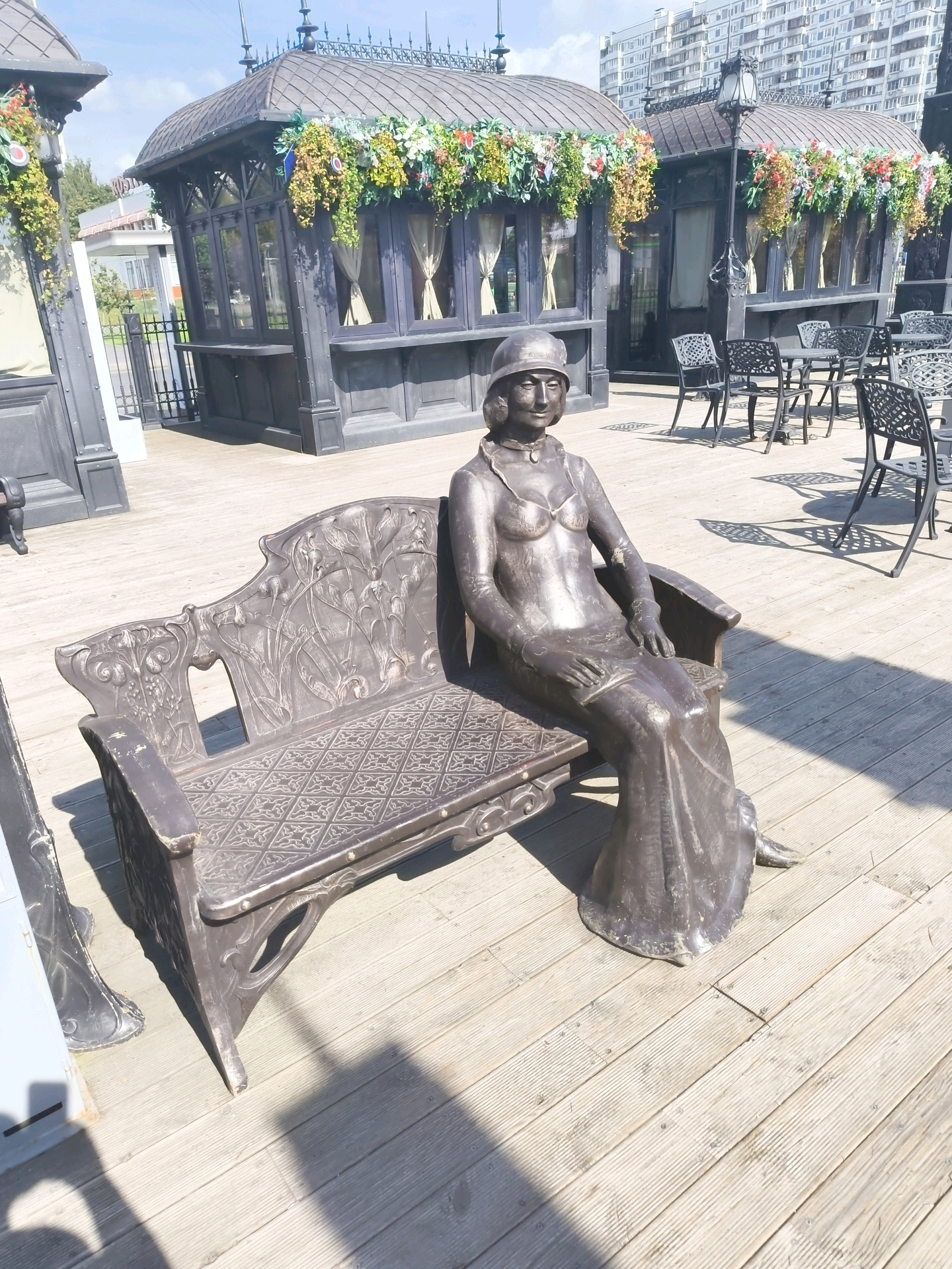 The image depicts a bronze statue of a woman seated on a decorative bench, positioned on a wooden deck. The statue is dressed in a long gown and a hat, with her hands resting in her lap. The bench itself is ornate, featuring intricate carvings and a patterned seat. In the background, there are several dark gazebos with decorative floral arrangements and outdoor seating areas, including tables and chairs. Beyond the gazebos, a modern residential building is visible. The scene is illuminated by sunlight, casting shadows on the wooden deck. This location is identified as the Sculpture Garden at the Krymskaya Embankment in Moscow, Russia.