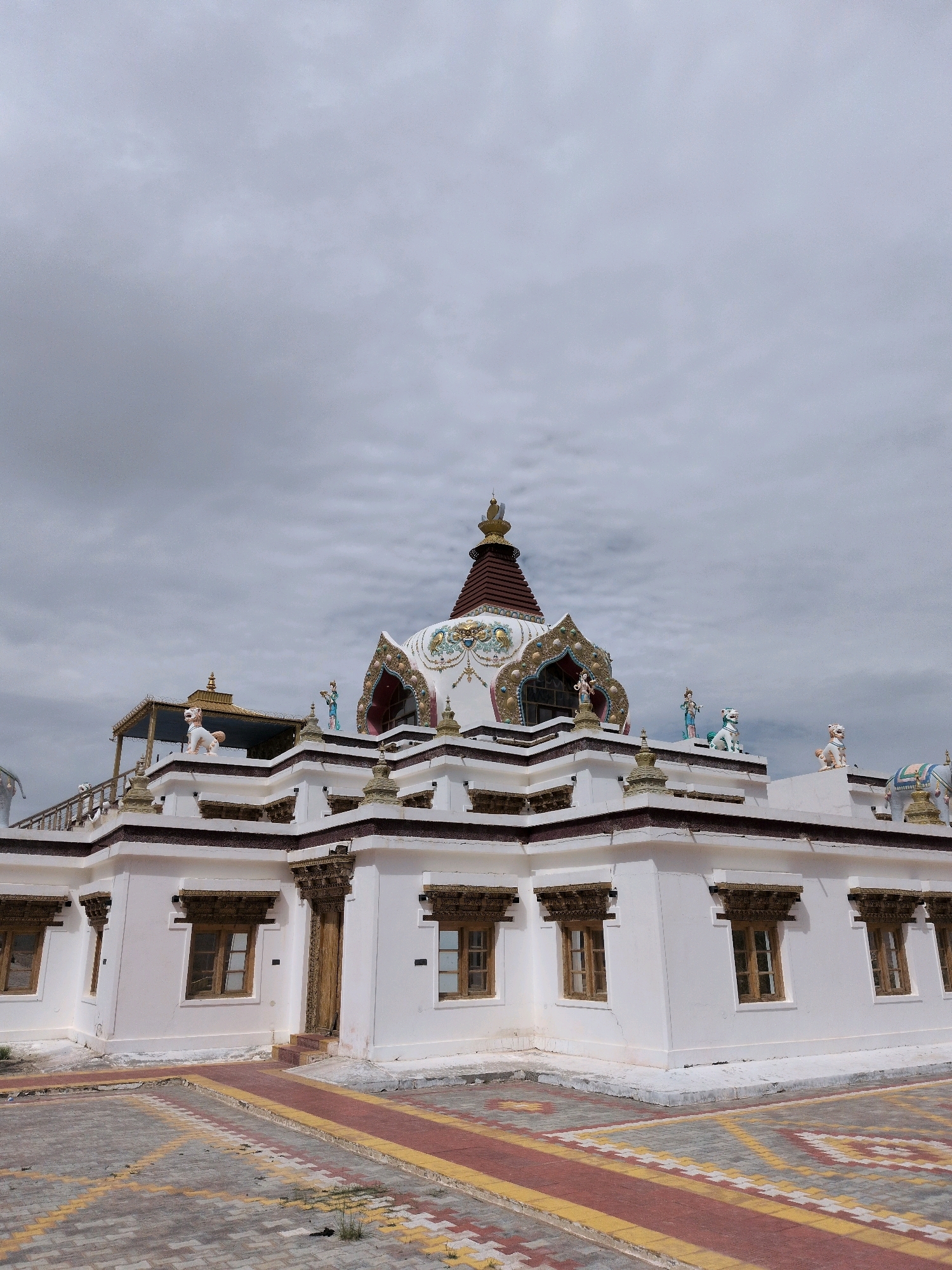 The image shows the exterior of a Buddhist stupa in Martselang, India, under a cloudy sky. The stupa is white with brown and gold decorative elements. It is adorned with statues of figures and animals on its upper levels. In the foreground, there is a paved area with red and yellow patterned tiles. The building has multiple windows with ornate wooden frames. The overall scene suggests a place of worship or a cultural landmark in a mountainous region.