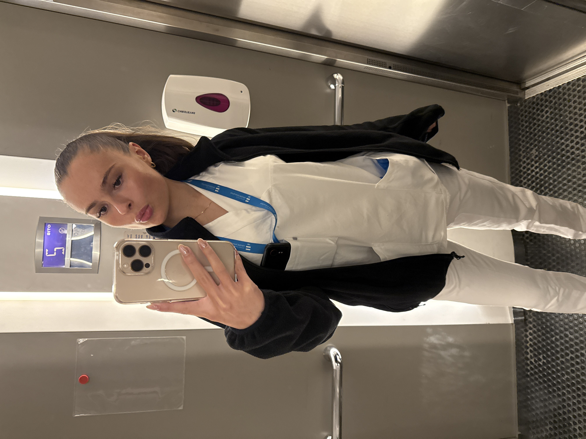 A young woman with blonde hair pulled back into a ponytail, wearing white scrubs and a black zip-up jacket, takes a selfie in an elevator. She is holding a gold iPhone in her right hand. A blue lanyard with a card is visible around her neck. A white dispenser is mounted on the wall to her right, and a digital display showing the number "5" is visible on the wall behind her. The elevator has a metal handle on the left wall and a textured metal floor. The overall environment appears to be an indoor, possibly institutional setting like a hospital or healthcare facility in Harstad, Norway.