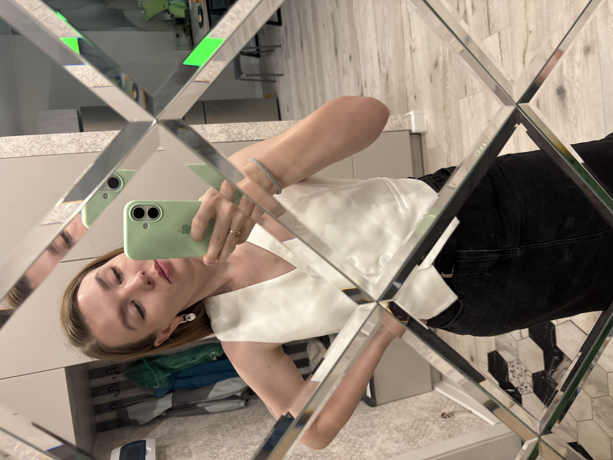A woman is taking a selfie in a diamond-patterned mirror. She is holding a green smartphone. The woman has blonde hair and is wearing a white top and black pants. The background includes a countertop, a tiled floor, and furniture. The city and location of Izhevsk, Russia are not discernible from the image.