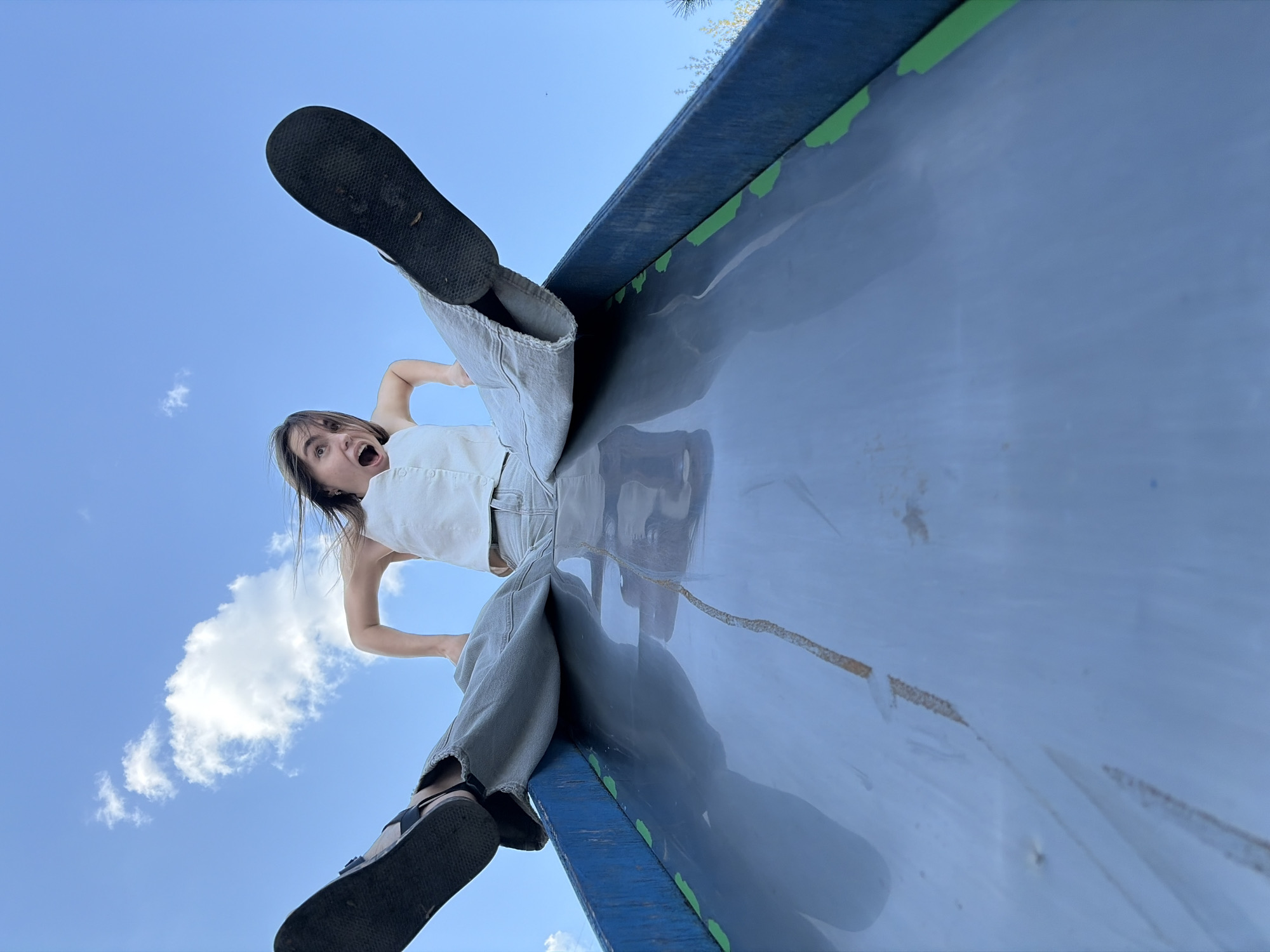 A young woman stands atop a blue and gray slide in Izhevsk, Russia. She wears a white top, gray pants, and dark sandals, with her bare feet facing upward. Her mouth is open in an expression of surprise or excitement. The slide is marked with green paint along the edges, and a clear blue sky with scattered clouds is visible in the background.