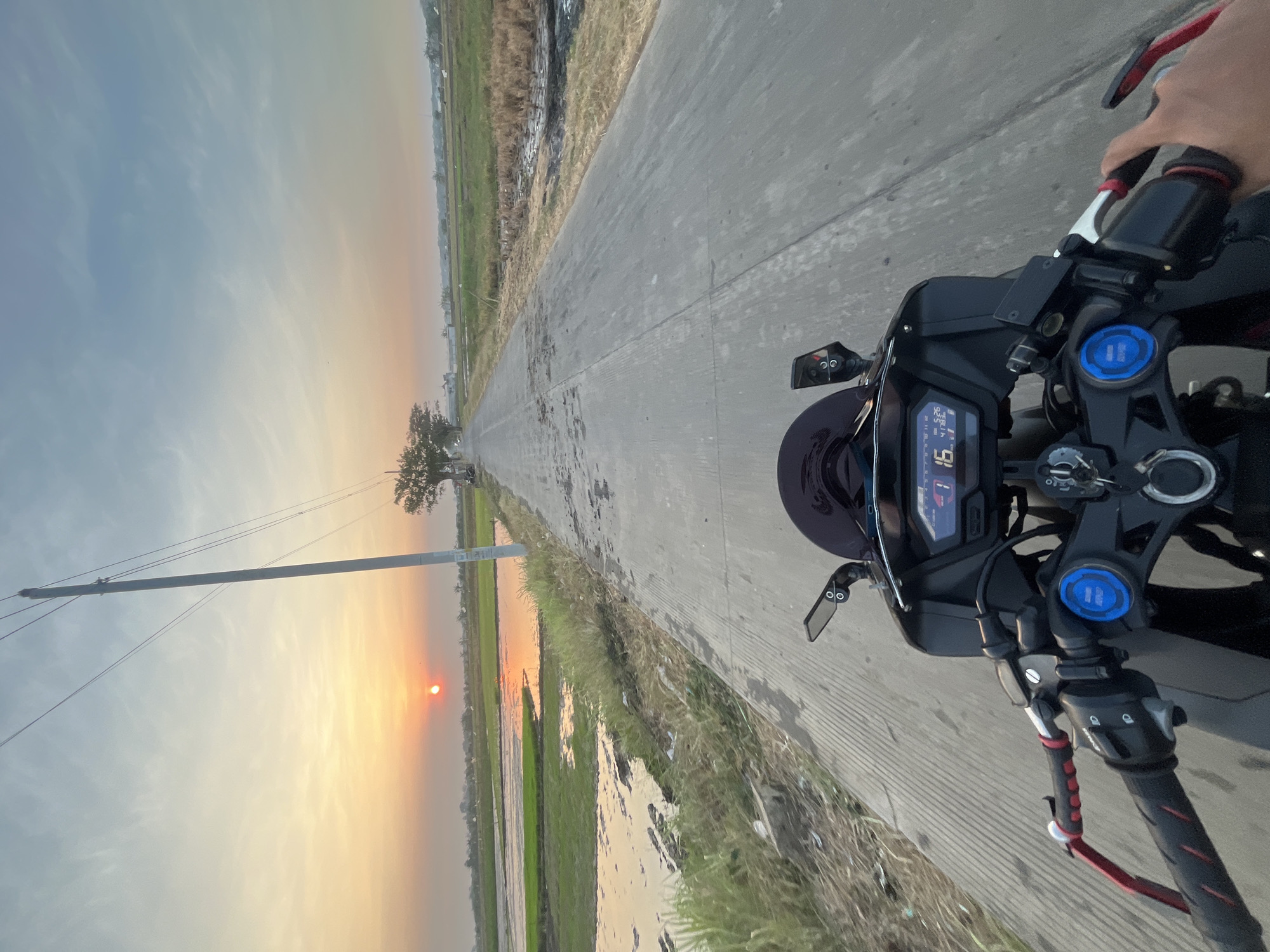 The image captures a first-person perspective from a motorcycle on a paved road in Pecangaan, Indonesia, at sunset. The sun, a vibrant orange orb, is low on the horizon, casting a warm glow across the sky and reflecting on the flooded rice fields to the left of the road. Tall grass and reeds line the edge of the road, and to the right, drier fields and sparse vegetation are visible. A lone, mature tree stands in the distance, near a cluster of buildings and more vegetation. Utility poles with wires run parallel to the road, indicating the presence of infrastructure. The motorcycle's handlebars, dashboard with a digital display showing "16" and other indicators, and mirrors are clearly visible in the foreground. A rider's hands are on the handlebars. The overall scene depicts a serene rural landscape during the golden hour.