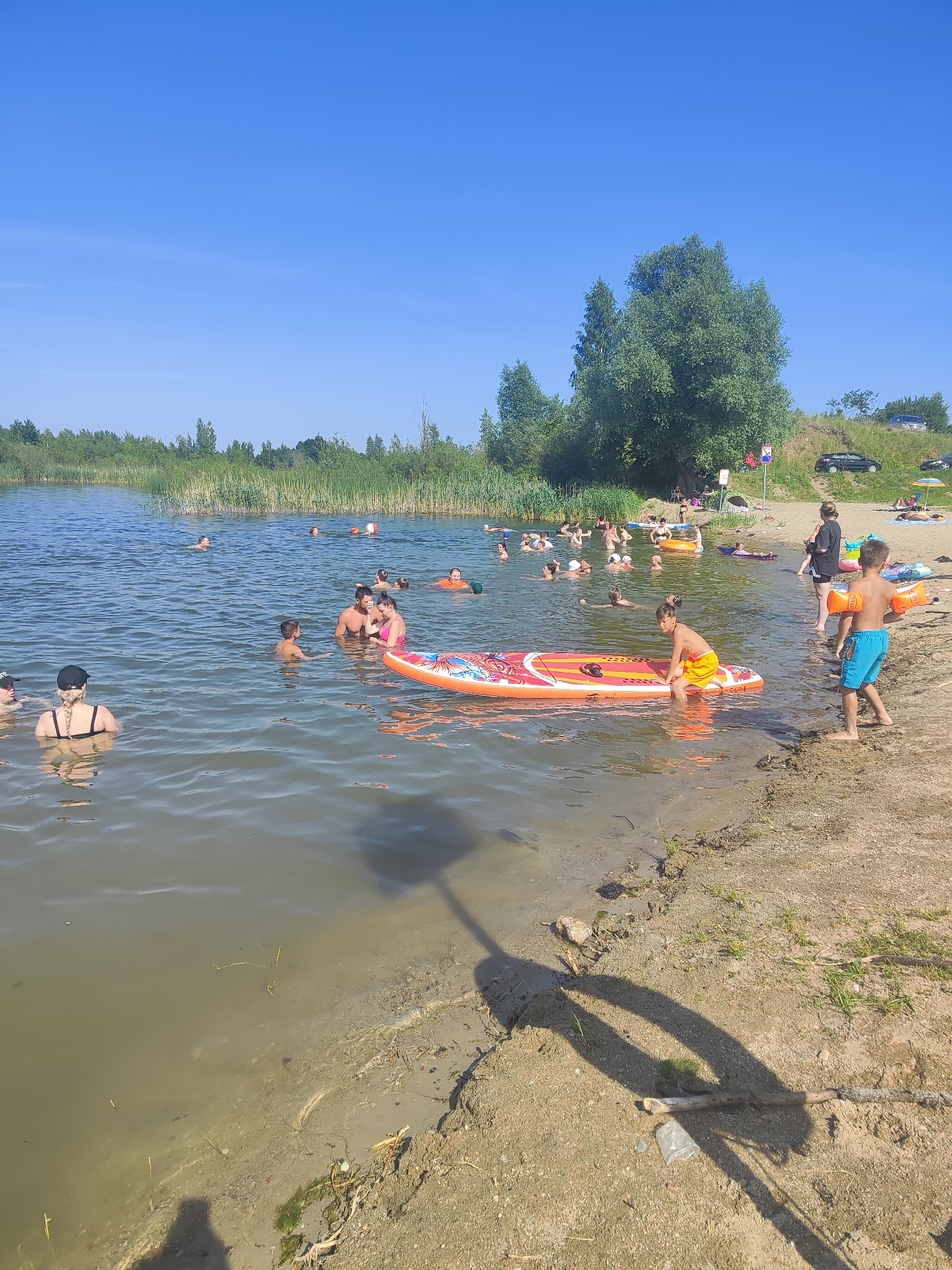A sunny day at a lake near Rovnoe, Russia, with many people enjoying the water and beach. Several individuals are swimming, wading, and playing in the lake. A large group is clustered in the water, some with children. One child is sitting on an orange and white inflatable stand-up paddleboard. Another young boy, wearing blue swim trunks and orange arm floaties, stands on the sandy shore. A woman in a black swimsuit and black cap is visible on the left side of the image, facing away from the camera. In the background, there's a line of trees and reeds along the lake's edge, with a gentle slope leading to a grassy area where several cars are parked. Two beach umbrellas are set up on the sand. The sky is clear and blue.