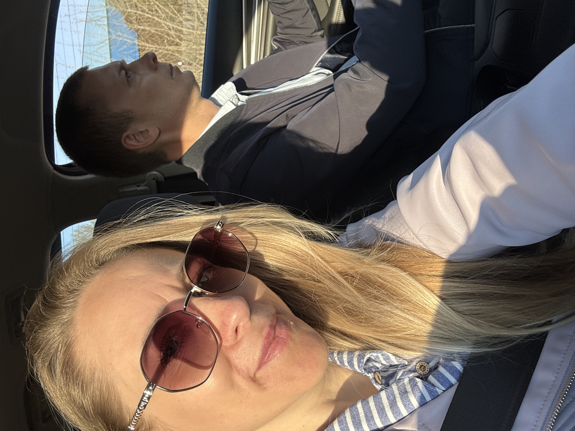 A selfie-style photo shows two people inside a vehicle. In the foreground, a blonde woman wearing sunglasses and a striped blue and white shirt. Behind her and partially out of focus is a man looking out the window on the passenger side. Based on the vehicle interior, it is daytime with clear skies outside. Location cannot be determined using the Chaikovsky reference.
