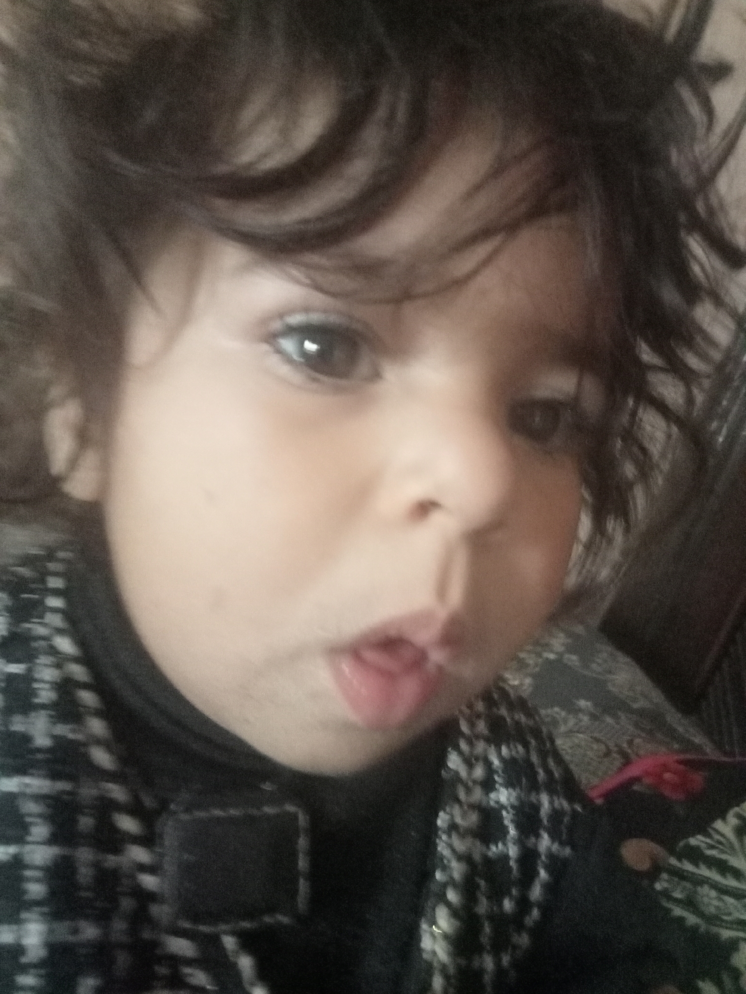 This image is a close-up, slightly blurred portrait of a young child, presumably taken indoors in Mansehra, Pakistan. The child, characterized by dark, wavy hair partially covering the forehead and cheeks, has light brown eyes looking directly at the camera. The mouth is open with the tongue slightly extended, suggesting a playful or pouting expression. The child is dressed in a dark-colored high-neck garment layered under an outer jacket or vest with a black and white checkered or woven pattern. A solid black rectangular patch is visible on the upper left portion of the outer garment. The background consists of a dark fabric with a subtle pattern, possibly paisley or floral motifs in shades of green and black, likely belonging to a cushion or blanket. A small, indistinct red object is partially visible in the lower right background. No specific architectural details are discernible to pinpoint an exact location within Mansehra beyond the general indoor setting.