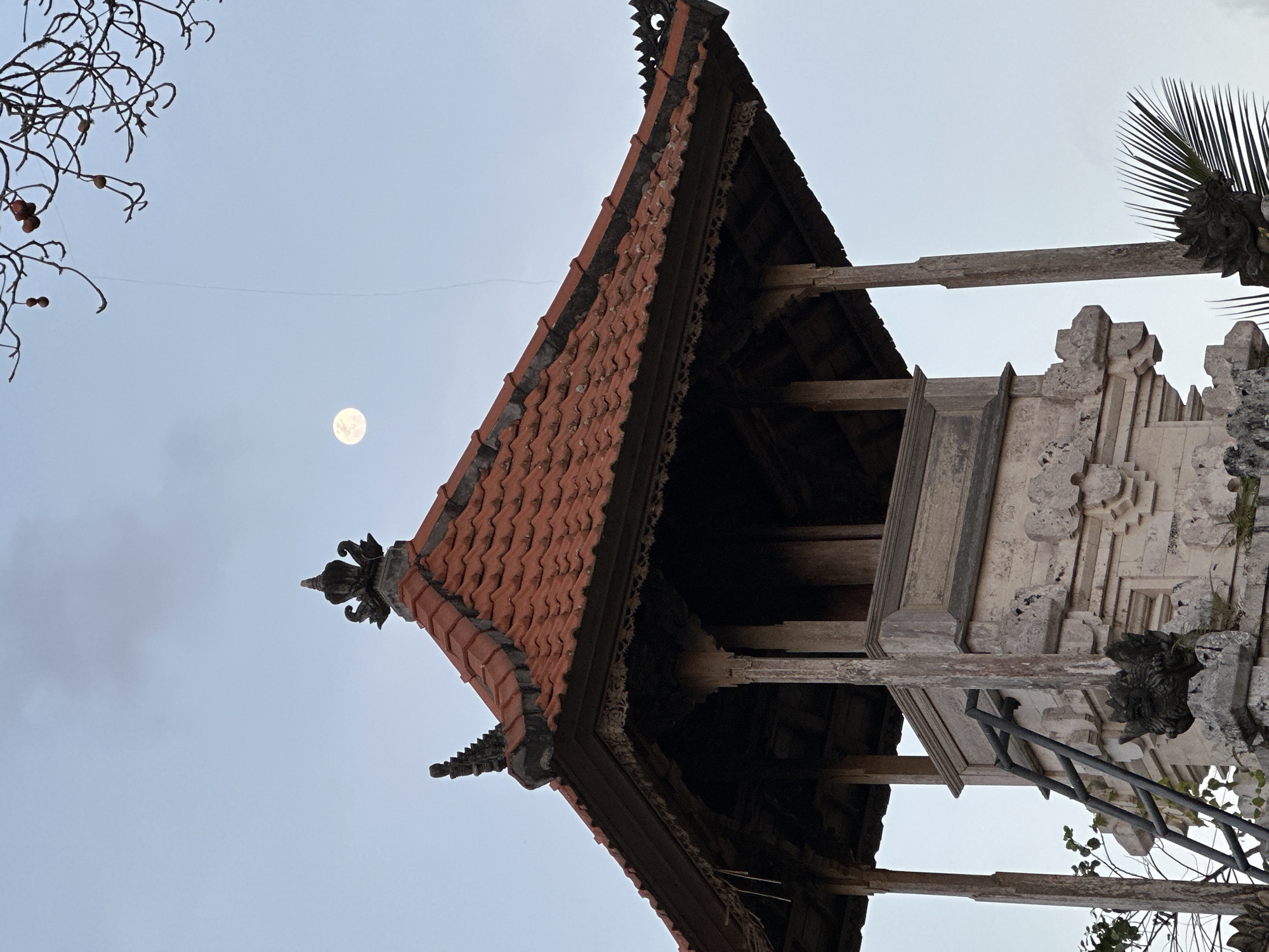 The image depicts an outdoor scene featuring a portion of a traditional building with a tiled roof, ornate wooden eaves, and stone carvings. A prominent, full moon is visible in the pale, overcast sky. To the left, bare tree branches with a few small, dark berries hang into the frame, suggesting a cooler climate or a dormant season, though the presence of palm fronds in the upper right indicates a tropical or subtropical setting. The architecture suggests a temple or a historically significant structure, common in Southeast Asian locations like Kuta Selatan, Indonesia. The lighting indicates it is either early morning or late evening, with the moon still clearly visible. There is no visible text in the image. The scene conveys a sense of tranquility and perhaps mystery, with the moon hanging in the sky above the ancient-looking structure.
