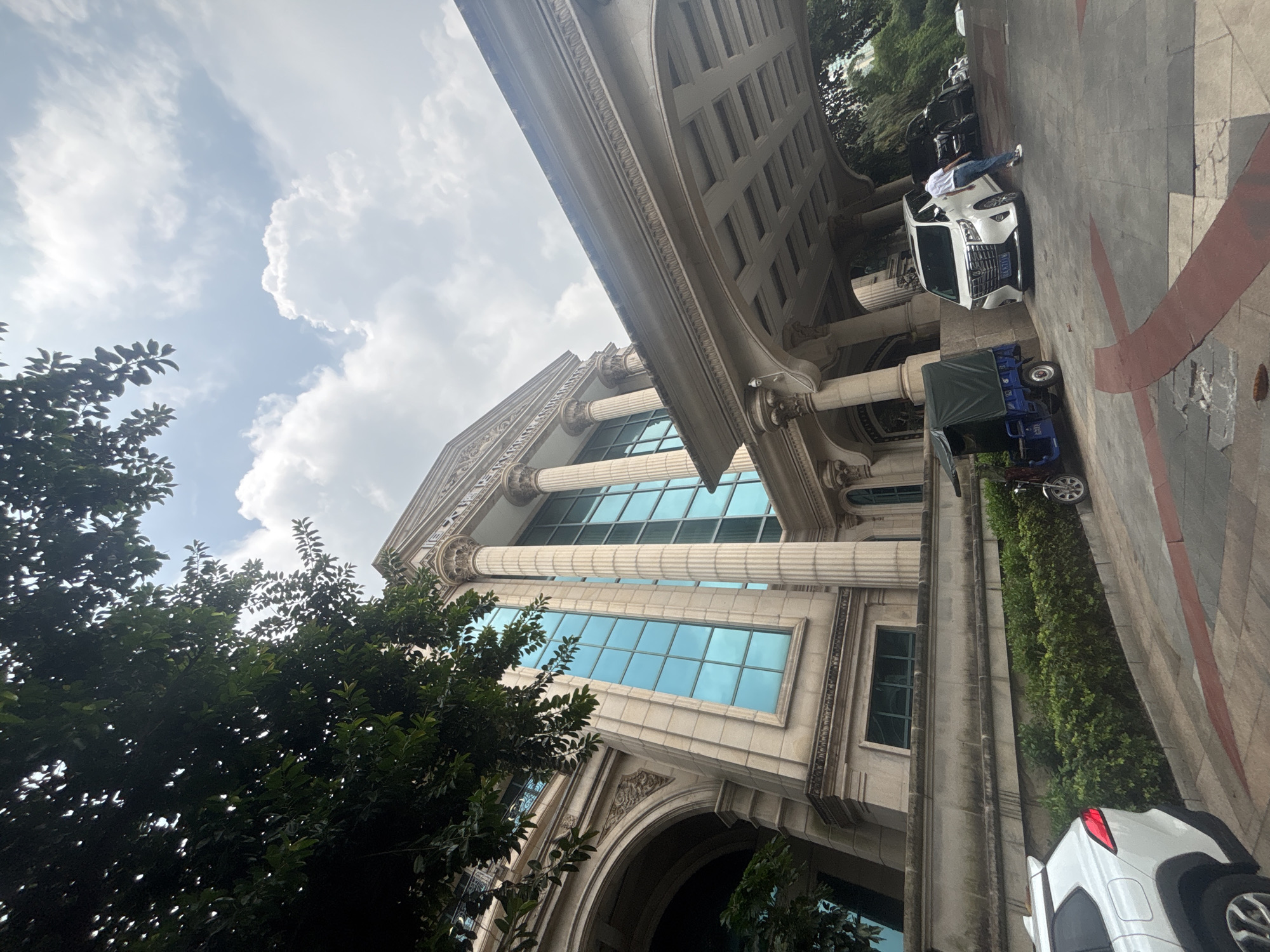 In Nanhai District, China, a low-angle shot captures the facade of a grand building, featuring classical architectural elements such as large columns and a prominent portico. The building's exterior is constructed from light-colored stone, and large, rectangular windows with a blue tint are visible. Lush green trees with visible leaves partially obscure the upper portions of the building and frame the sky, which is filled with scattered clouds.
In the foreground, a white SUV is parked on a paved area. To the left of the SUV, a blue three-wheeled vehicle with a dark green canopy is parked. A person, wearing a white t-shirt and blue jeans, is standing near the white SUV, appearing to interact with it or step out of it. The ground is a patterned pavement with red and gray sections, some of which show signs of wear and tear. The overall scene suggests a public or commercial space, possibly an entrance to a hotel or a large establishment, under an overcast sky.