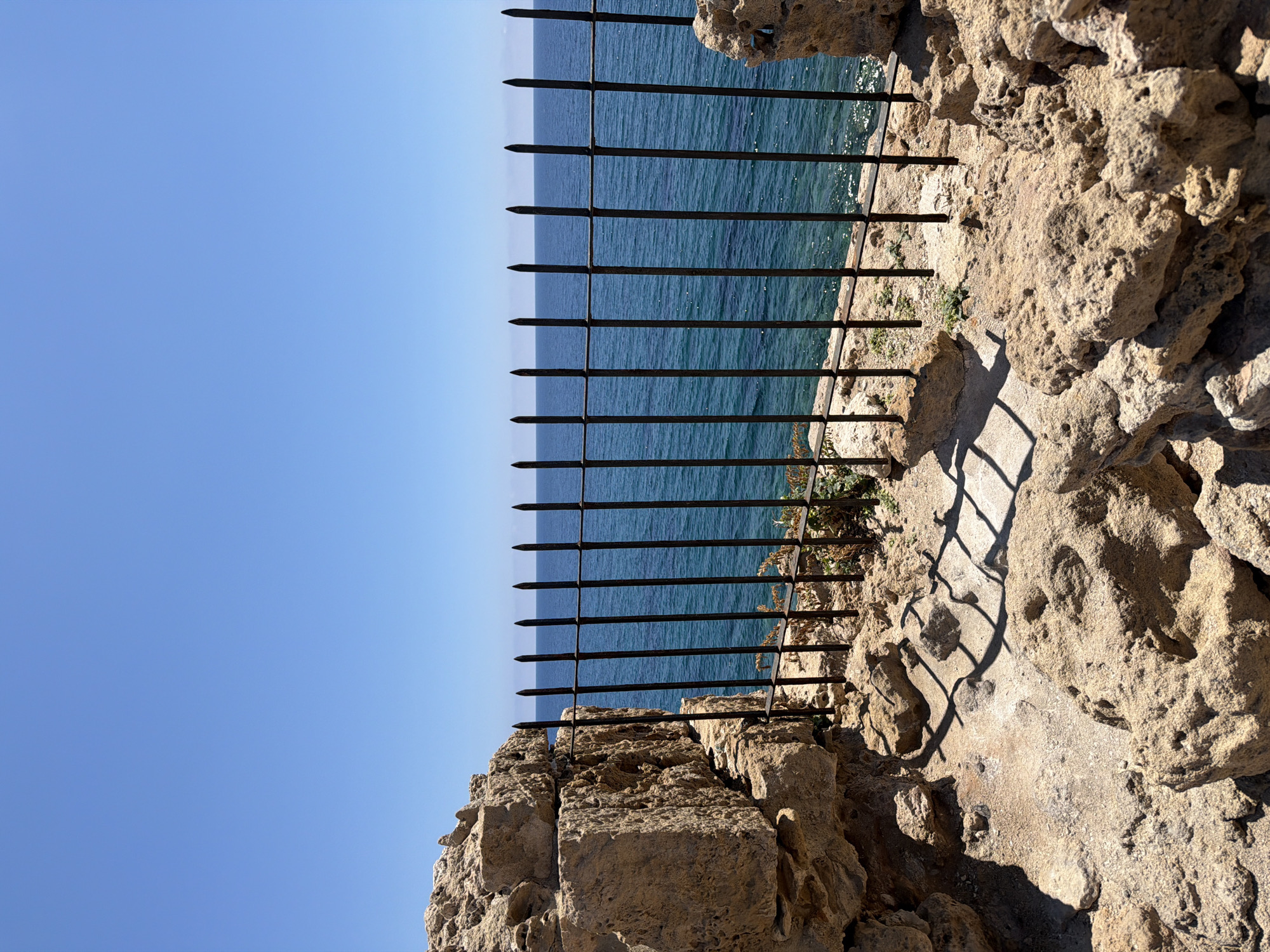 The image captures a scene in Paphos Municipality, Cyprus, showcasing a rocky cliff overlooking the sea. A black wrought-iron fence, with pointed tips, runs along the edge of the cliff, acting as a safety barrier. The clear blue sky contrasts with the textured, tan-colored rock formations. The sea, visible through the fence, appears calm with gentle ripples. The shadows cast by the fence on the rocks suggest a sunny day. There are no people visible in the scene.