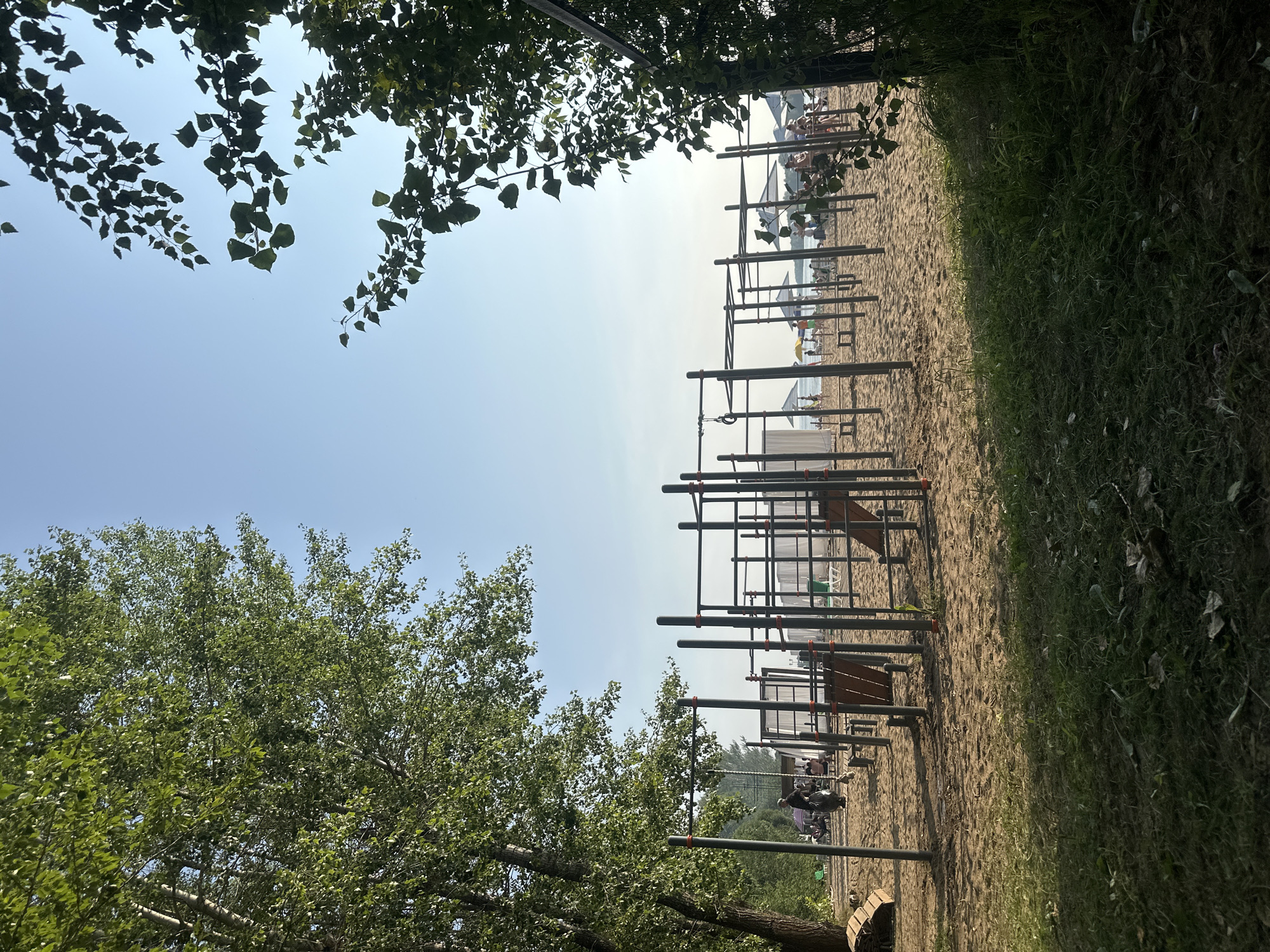 A low-angle shot captures a sandy outdoor gym area with various workout equipment, including pull-up bars and parallel bars, on a beach. The ground is a mix of sand and grass, with trees framing the scene. In the background, the beach is populated with people relaxing under umbrellas and near the water. The sky is clear and blue, suggesting a warm day. This location is identified as the beach at Ust-Kachka, Russia.