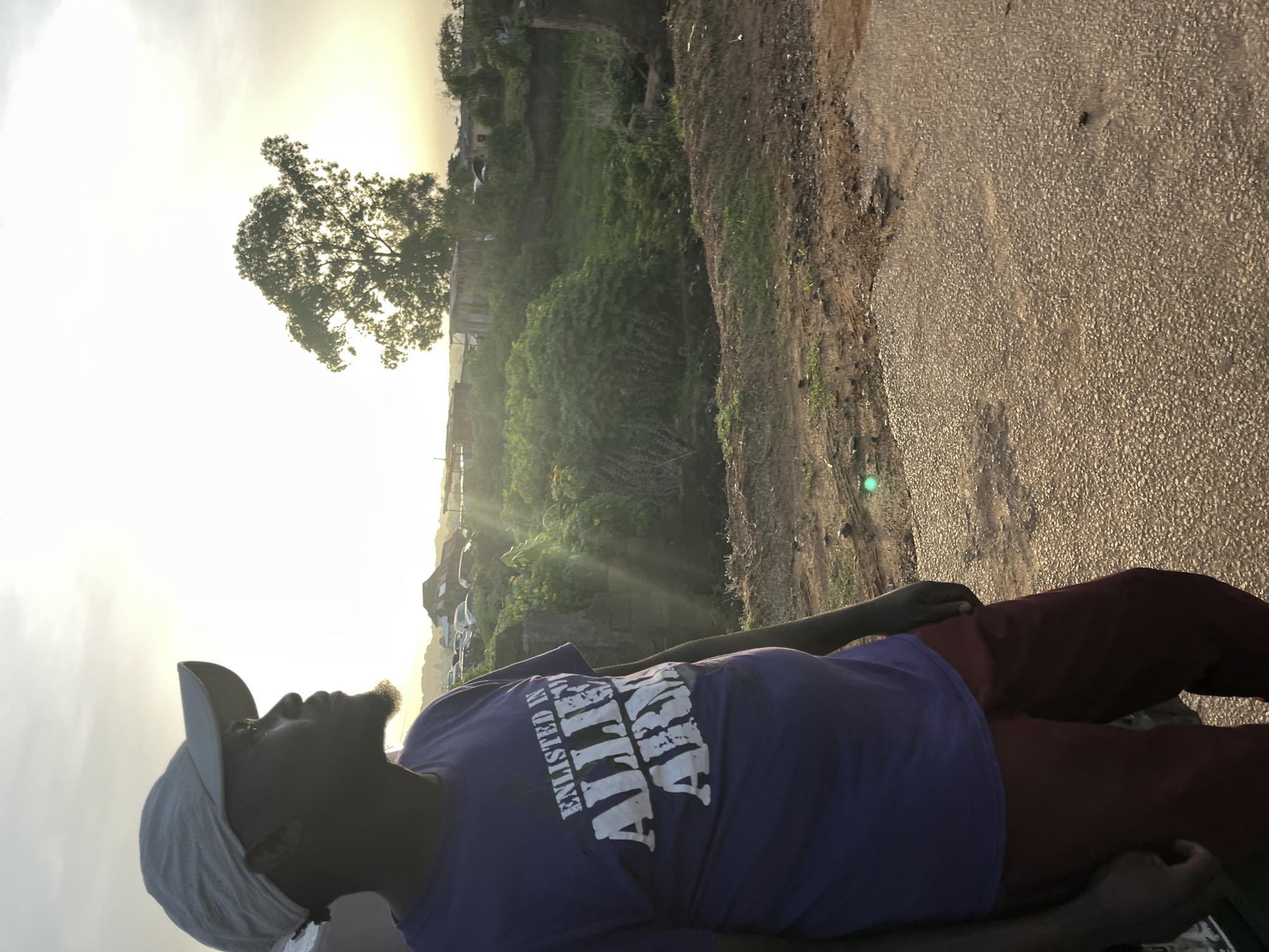 The media file features a person in the foreground, seen in profile and looking upwards towards a bright, golden sky. The person appears to be male, wearing a purple t-shirt, dark red or maroon trousers, and a light-colored baseball cap worn backward. The text "ENLISTED IN ALLIE ARMY" (or similar, partially obscured) is visible on the front of the t-shirt.
The scene is set outdoors, likely during sunrise or sunset, characterized by strong backlighting and lens flares from the sun, which is positioned in the upper left of the frame. This lighting creates a silhouette effect on the person and a large, full tree located in the mid-ground. Beyond the person and the immediate foreground, there is a rough, textured ground that appears to be an unpaved road or path. Further back, dense green foliage and smaller trees are visible, with some distant buildings nestled beneath the hazy, bright sky. The setting suggests a rural or semi-urban environment in Rafin Par, Nigeria. The person is standing still, observing the sky or the sun, giving a contemplative or appreciative impression.