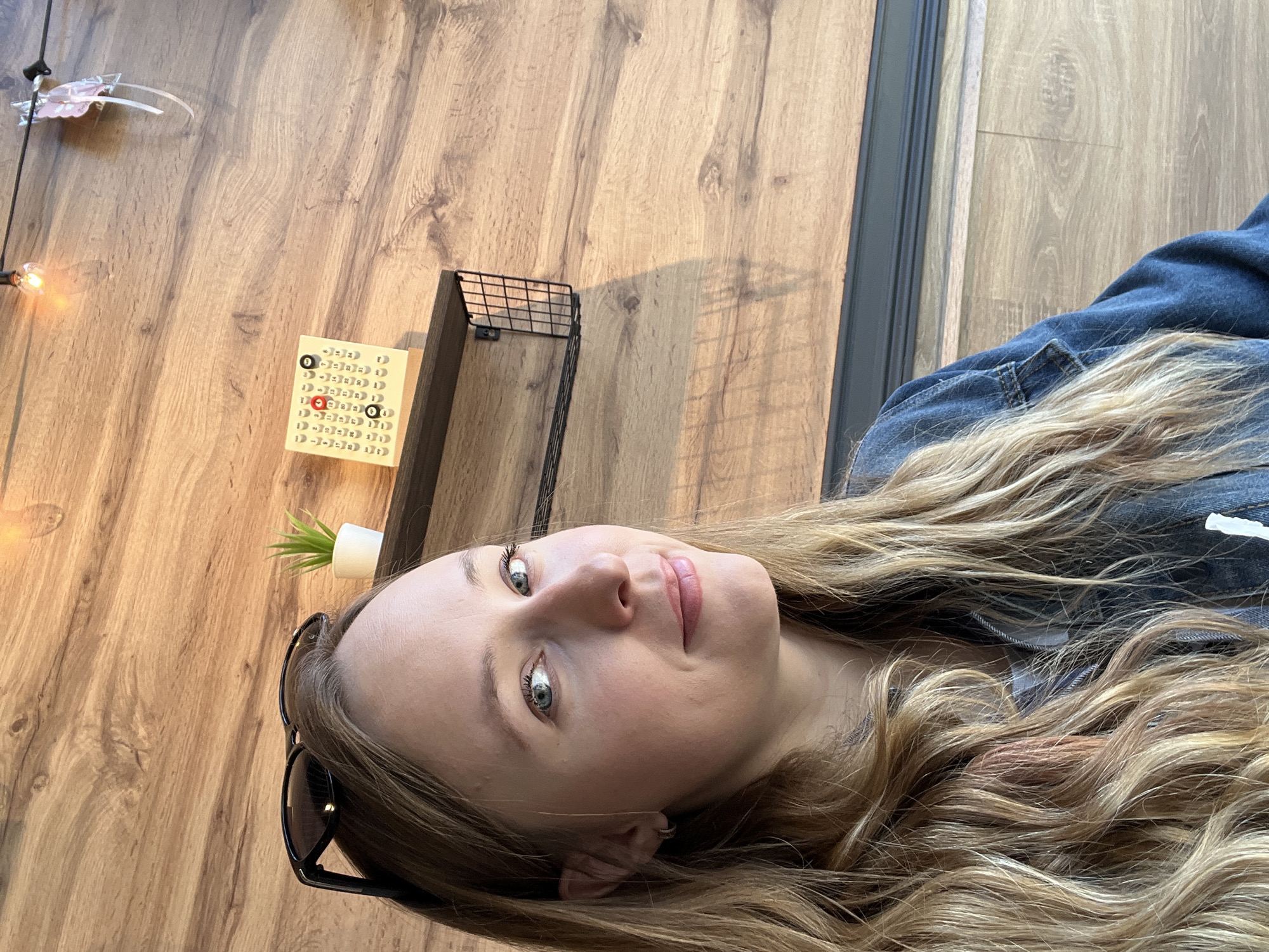 A medium shot depicts a light-skinned female with long blonde and brown hair. She is wearing a dark blue denim jacket and has light blue eyes. To the left of the frame, there is a faux wood-grain wall. A small calendar with red numbers hangs on the wall, along with a small plant in a white pot, and a wire basket. A pair of sunglasses is perched on top of her head. The location and presence in Mytishchi, Russia, are unknown.