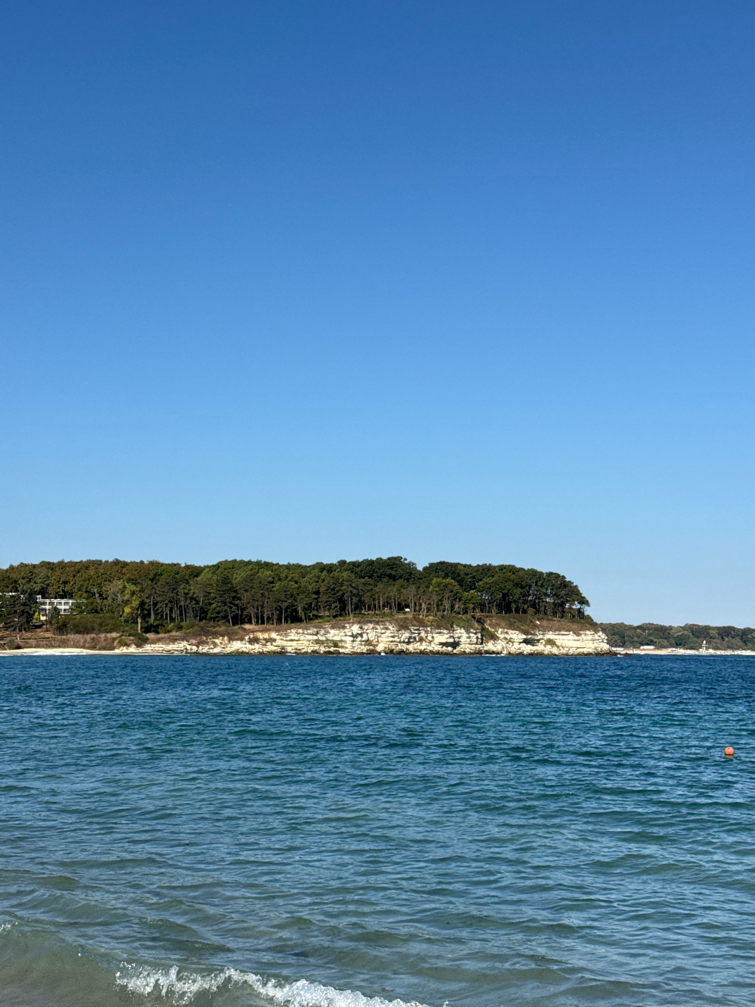 The image depicts a coastal landscape in Kiten, Bulgaria, under a clear blue sky. In the foreground, the blue sea gently laps at a sandy shore, with small waves breaking. A distinct orange buoy is visible in the middle distance of the water.
The middle ground is dominated by a white, rocky cliff face that extends horizontally across the frame. This cliff is topped by a dense, green forest, with numerous trees creating a thick canopy. On the left side of the cliff, a modern white building with multiple windows is partially visible, nestled amongst the trees. The shoreline continues to the right, with more trees and vegetation along the coast.
The background is a vast expanse of clear, cloudless blue sky, providing a simple backdrop to the scene. The overall impression is one of a serene natural setting by the sea.