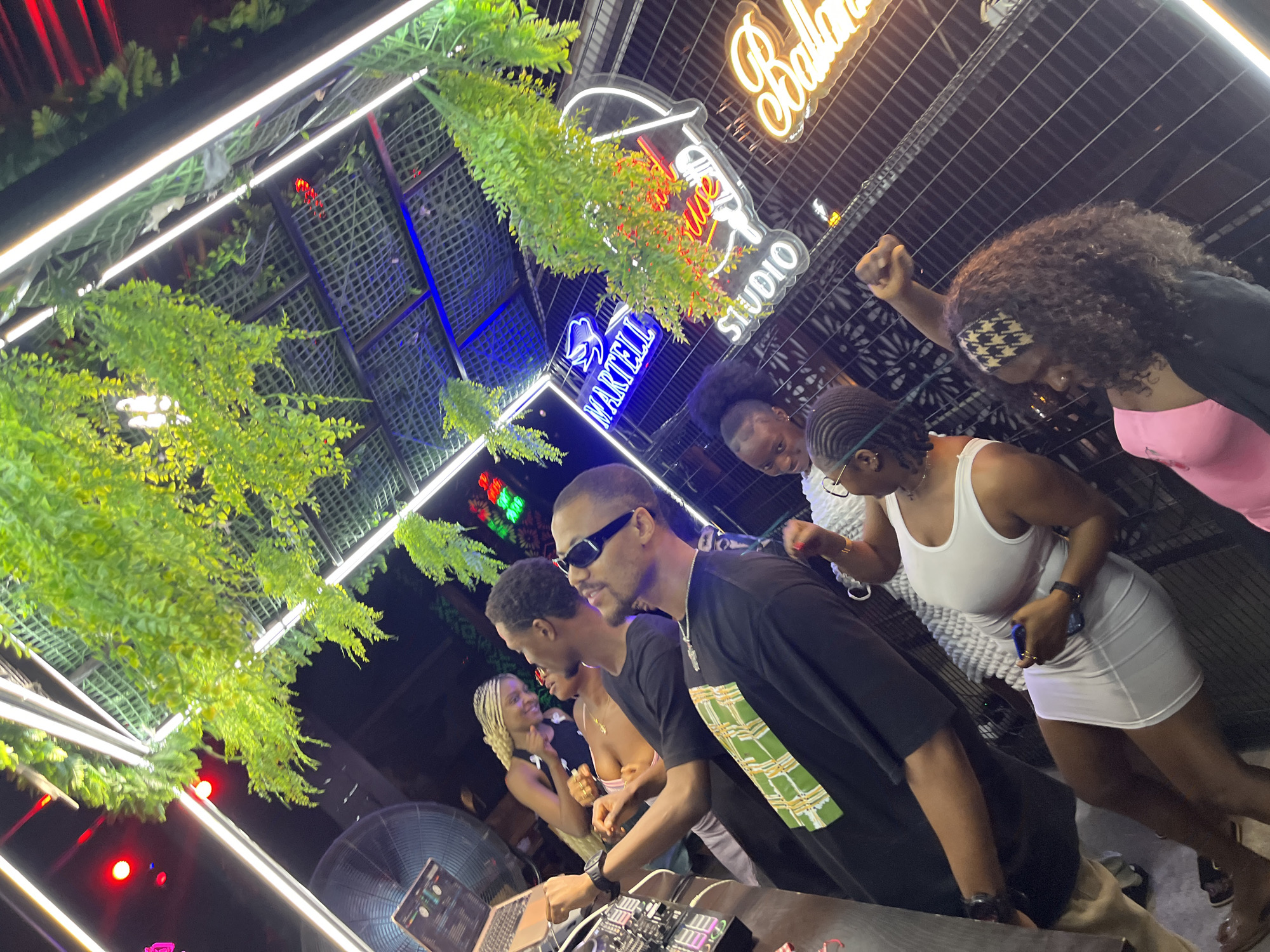 The image depicts an outdoor nighttime scene at a venue in Howayi, Nigeria. The setting appears to be a rooftop or elevated area, with neon lights and hanging artificial plants creating a vibrant atmosphere. In the foreground, a group of young adults are socializing. One individual, wearing sunglasses and a black t-shirt with a green and white checkered design, is facing the camera and smiling. Another person, dressed in a white tank top and a short white skirt, is also facing the camera and holding a phone. To their left, a person in a black tank top with blonde braided hair is looking towards the foreground. In the background, several neon signs are visible, including one that reads "MARTELL STUDIO" and another with the word "Balloo" in cursive. The overall impression is of a lively social gathering.